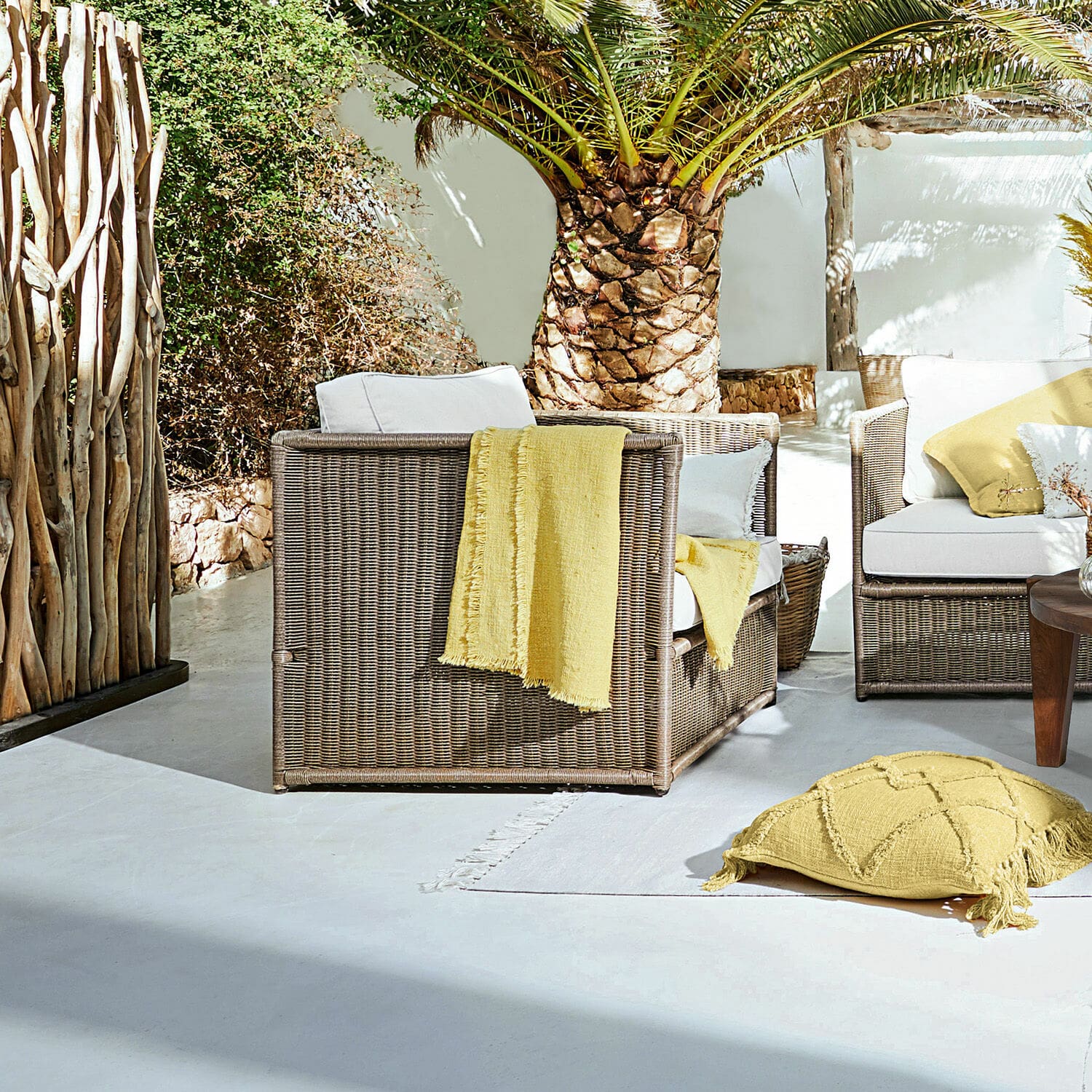 Sillón Lesmont Conjunto de muebles de jardín de ratán marrón con cojines blancos y detalles amarillos en terraza soleada
