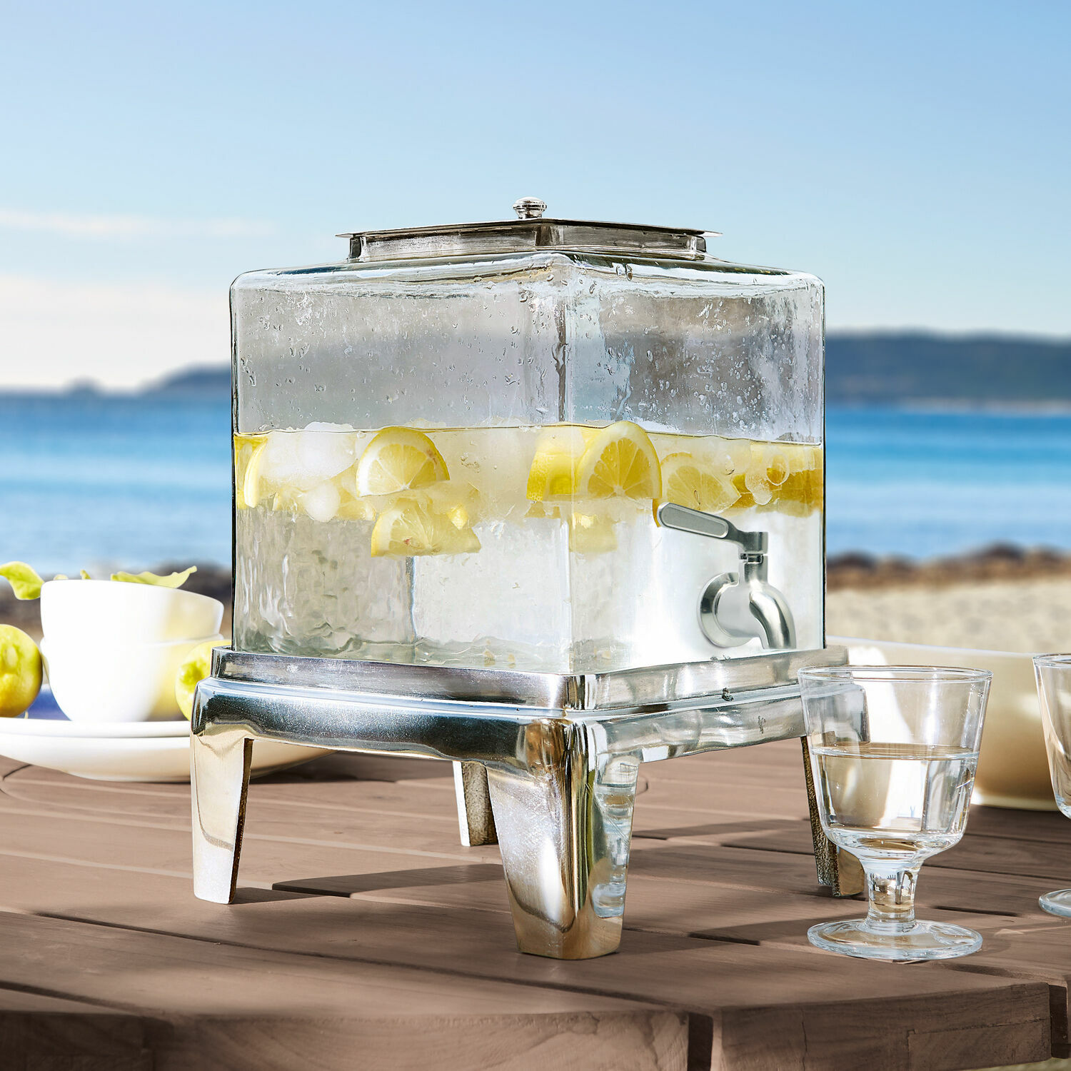 Dispensador de bebidas Osric Dispensador de bebidas de vidrio con base metálica y grifo, lleno de agua con limón y hielo