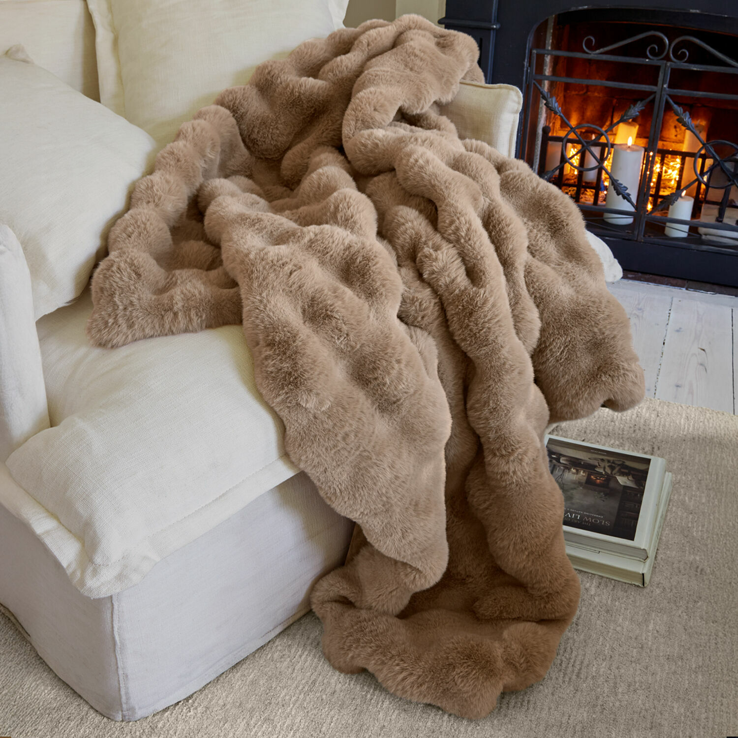 Plaid Zalina Manta beige de piel sintética sobre sofá claro junto a chimenea encendida para ambiente acogedor