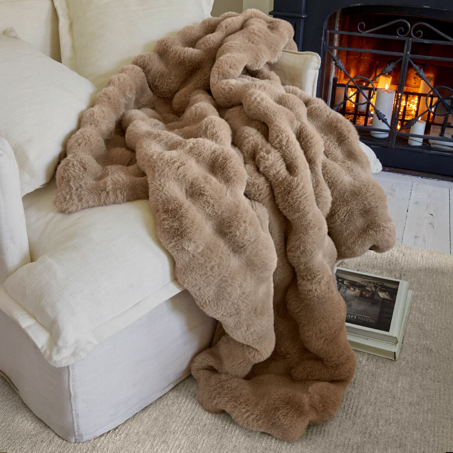Plaid Zalina Manta beige de piel sintética sobre sofá claro junto a chimenea encendida para ambiente acogedor