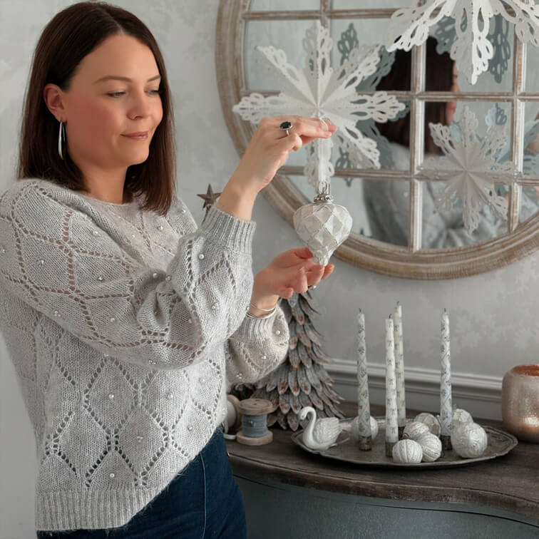 Mujer con suéter gris con perlas sostiene adorno navideño blanco en interior acogedor con decoración invernal