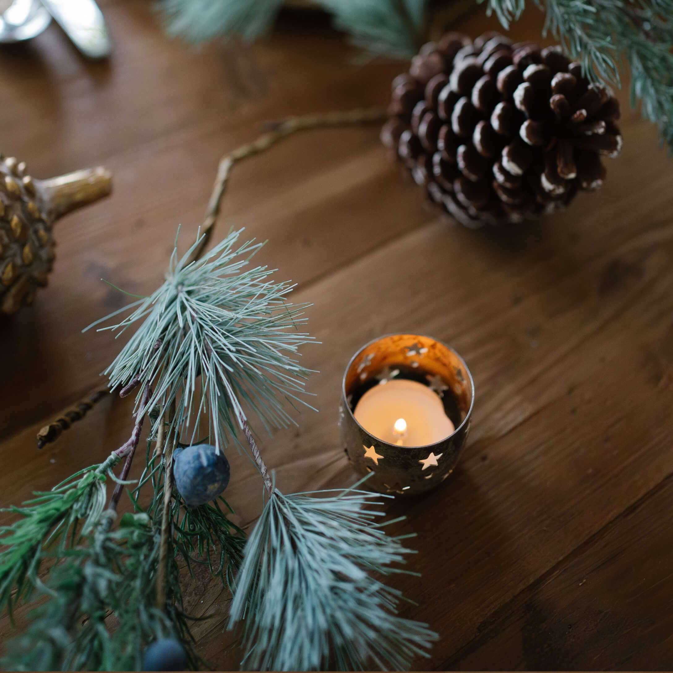 Portavelas metálico con estrellas recortadas y vela encendida sobre mesa de madera con decoración de piñas y ramas