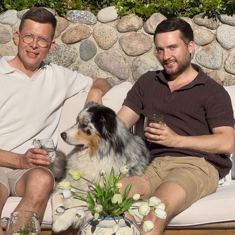 Dos hombres sentados en un sofá exterior con un perro pastor australiano y un jarrón con tulipanes blancos.