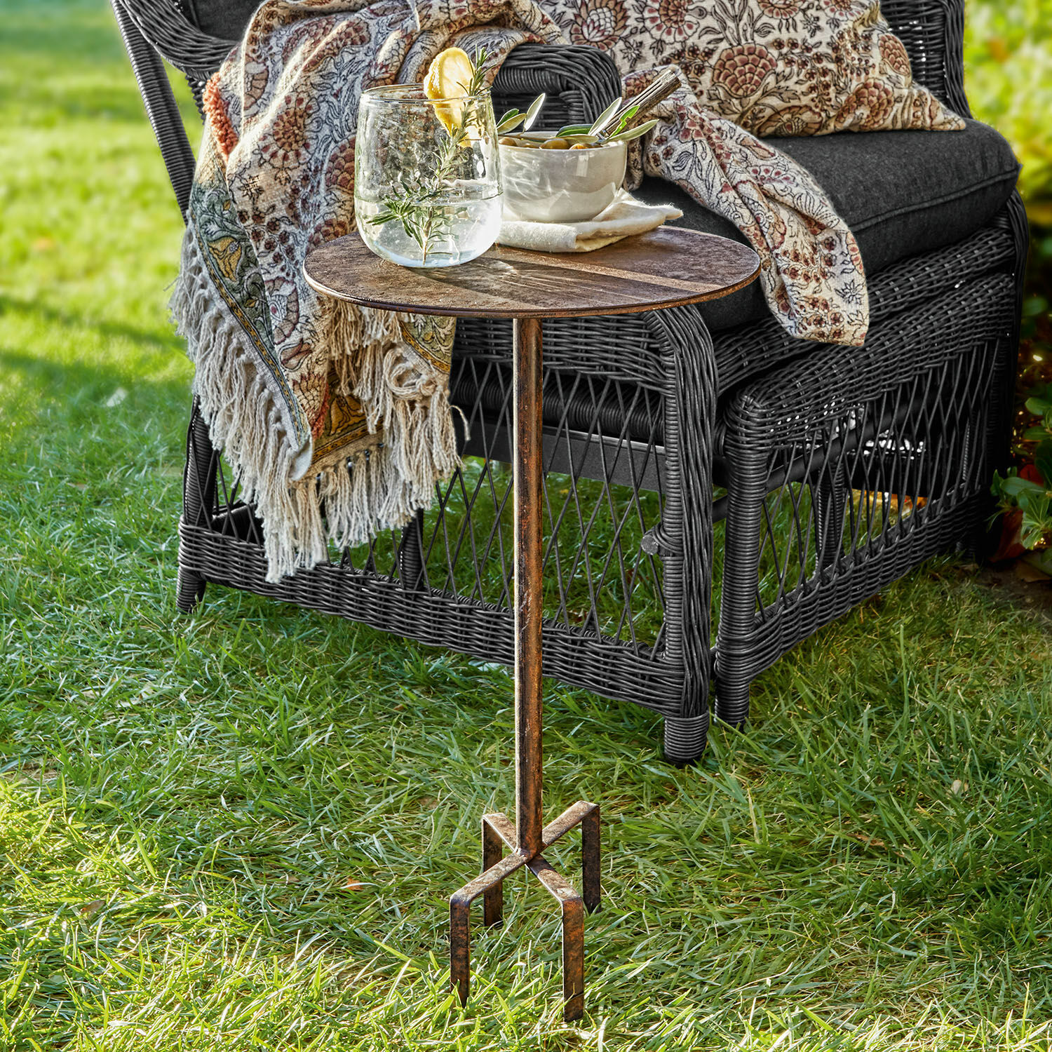 Mesa pequeña Fermont Mesa auxiliar de metal con pátina vintage sobre césped junto a sillón de mimbre con cojín y manta