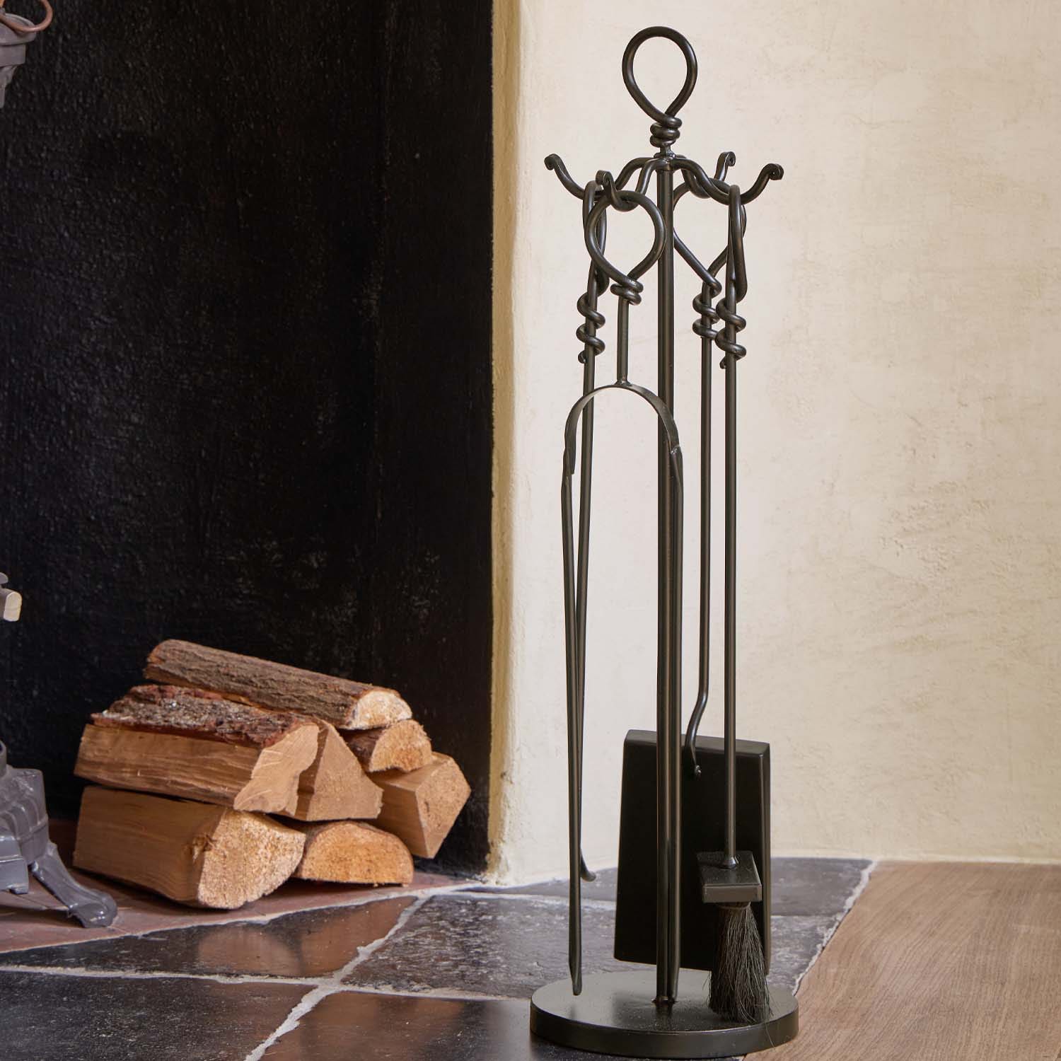 Conjunto de herramientas para chimenea de metal negro junto a leña apilada sobre suelo de piedra