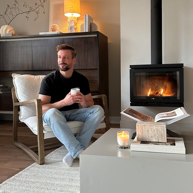Hombre sentado en sillón de madera con cojín blanco, sostiene taza, chimenea y vela en mesa de salón