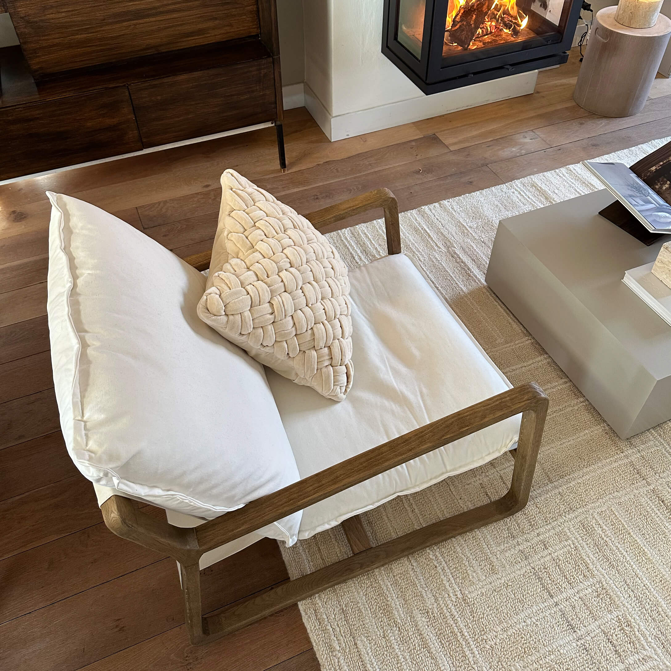 Sillón con estructura de madera y cojines crema sobre alfombra clara junto a chimenea en sala de estar
