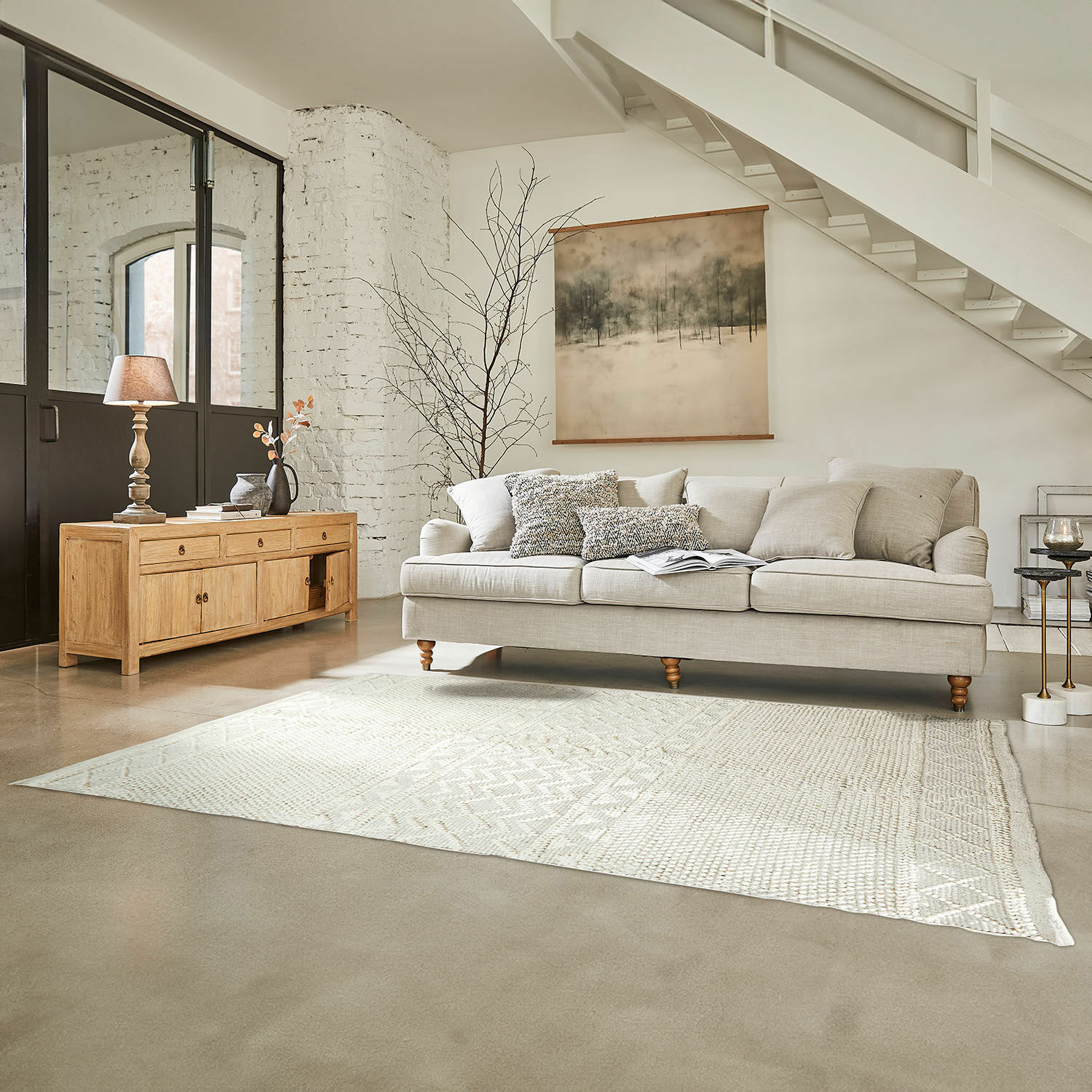 Sofá beige con cojines y aparador de madera clara en salón luminoso con alfombra y decoración de pared