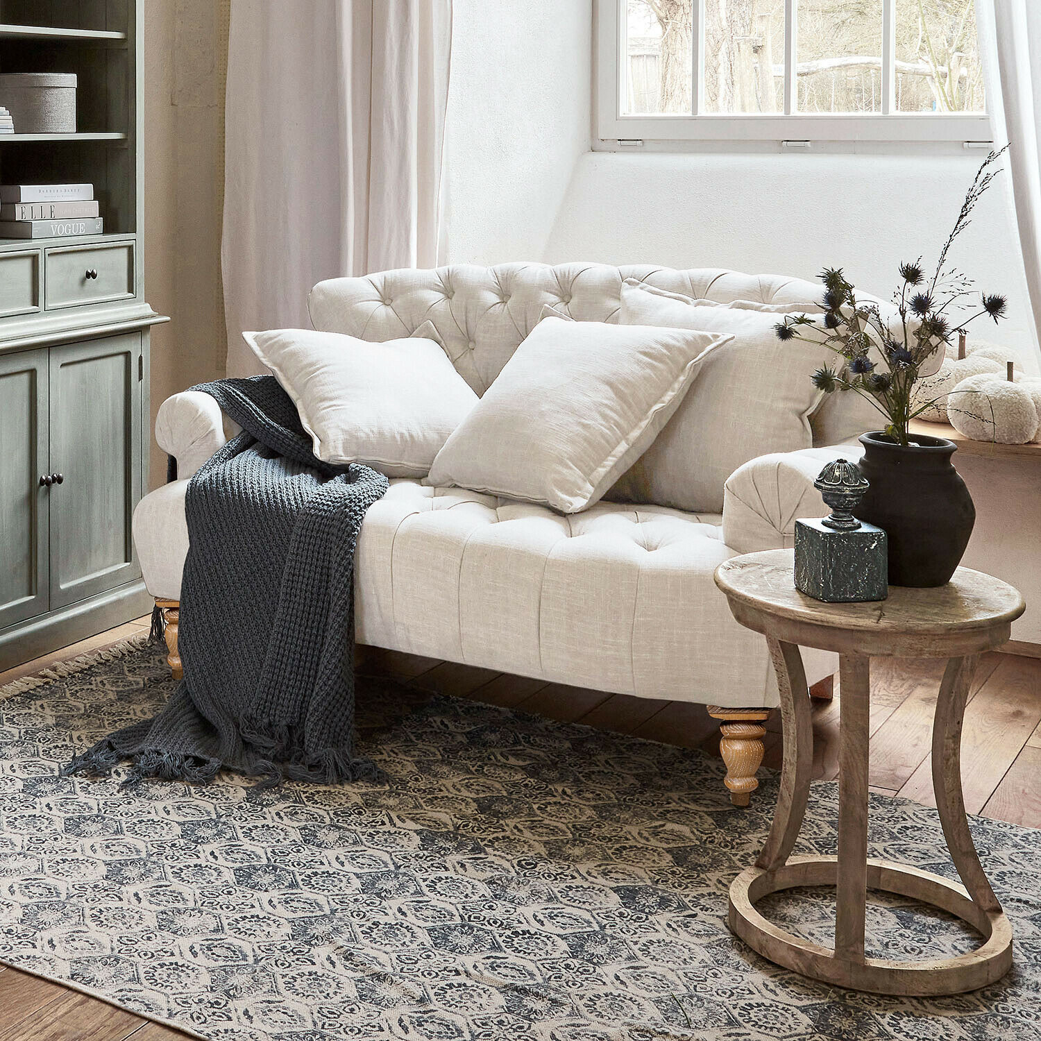 Sofá Springfield Village Sofá beige capitoné de lino con cojines y manta gris de punto en salón acogedor con mesa de madera