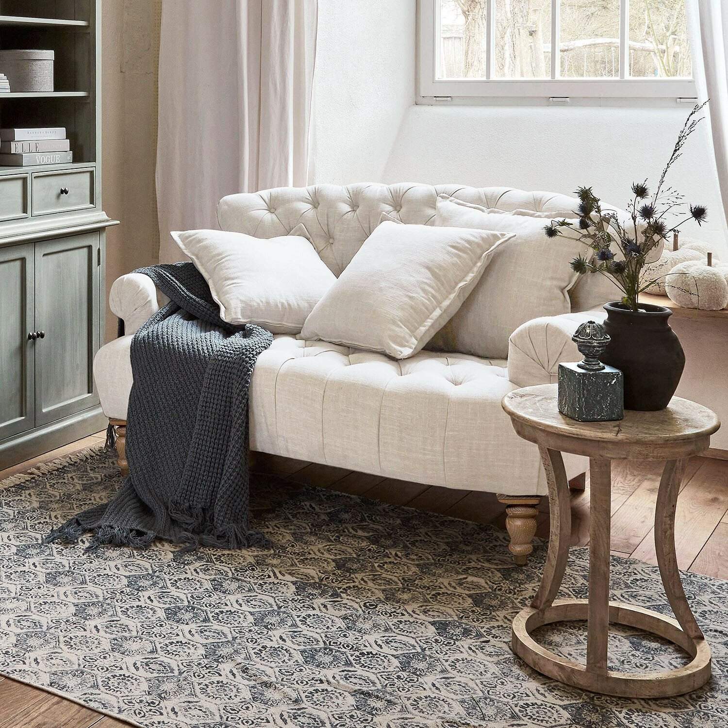 Sofá Springfield Village Sofá beige capitoné de lino con cojines y manta gris de punto en salón acogedor con mesa de madera
