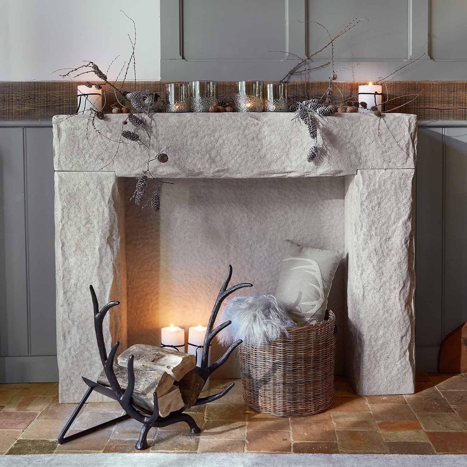 Chimenea de piedra blanca con ramas y velas decorativas, cesta de mimbre con cojín y manta de piel