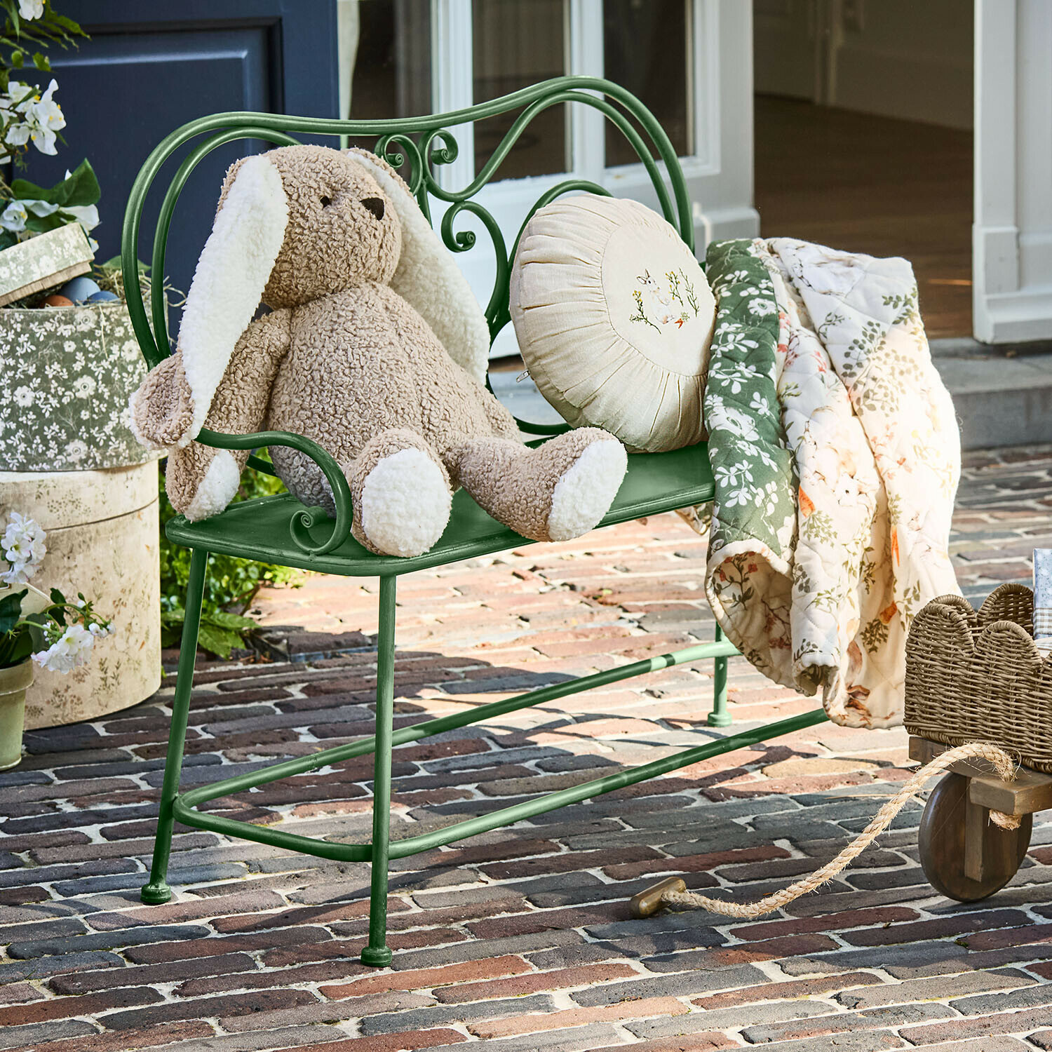 Banco Fernwick Banco metálico verde con peluche de conejo beige y manta floral sobre patio de ladrillo