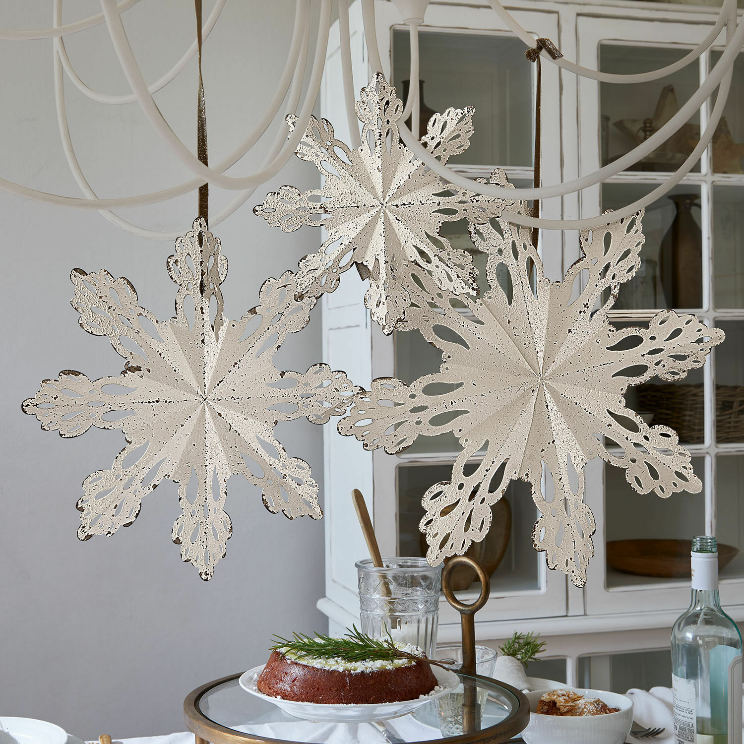 Set de 3 decoraciones de pared Lirvaya Copos de nieve decorativos de metal color crema colgados en habitación luminosa con pastel sobre mesa