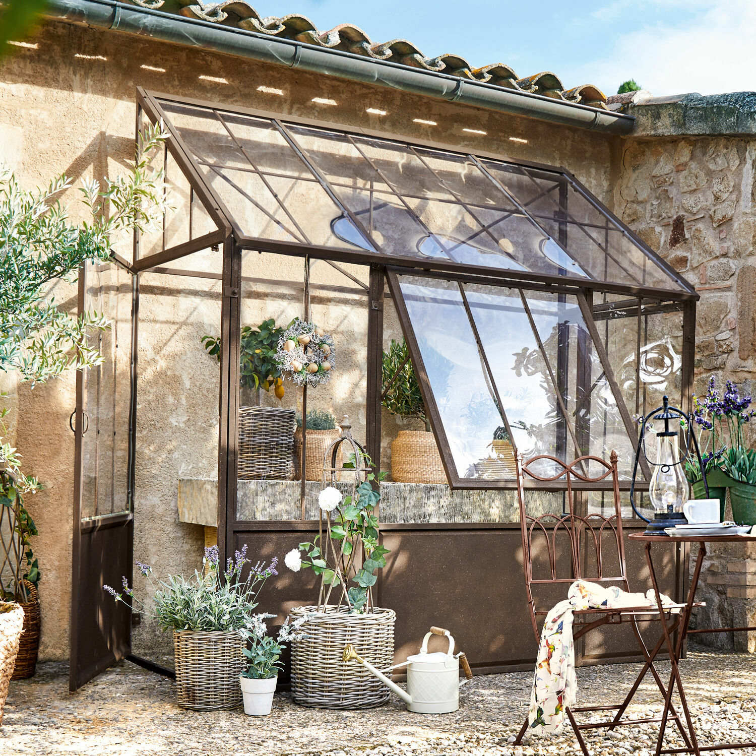 Invernadero Essone Invernadero de jardín con estructura metálica marrón, plantas y muebles de jardín en exterior soleado