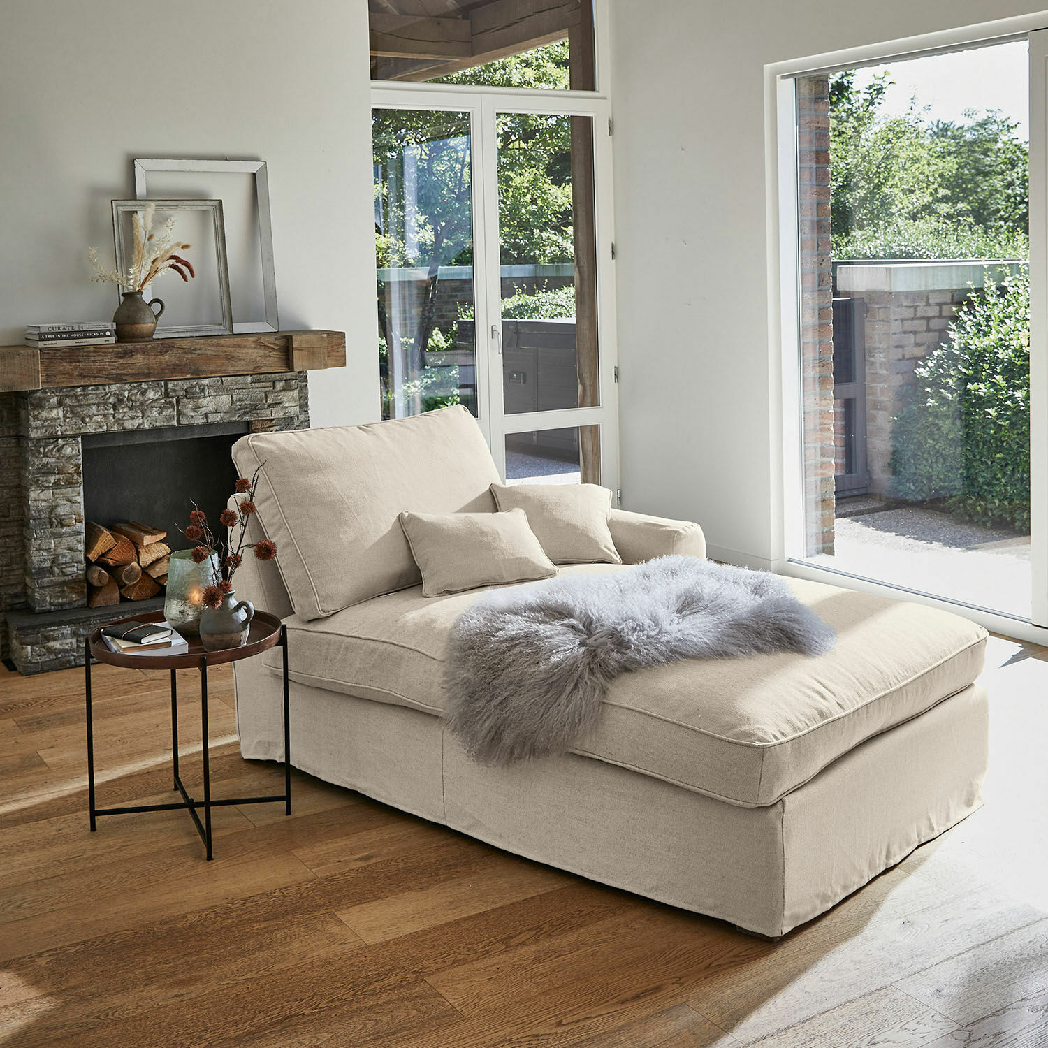 Chaise longue beige de tela con cojines y manta gris peluda en sala luminosa con chimenea