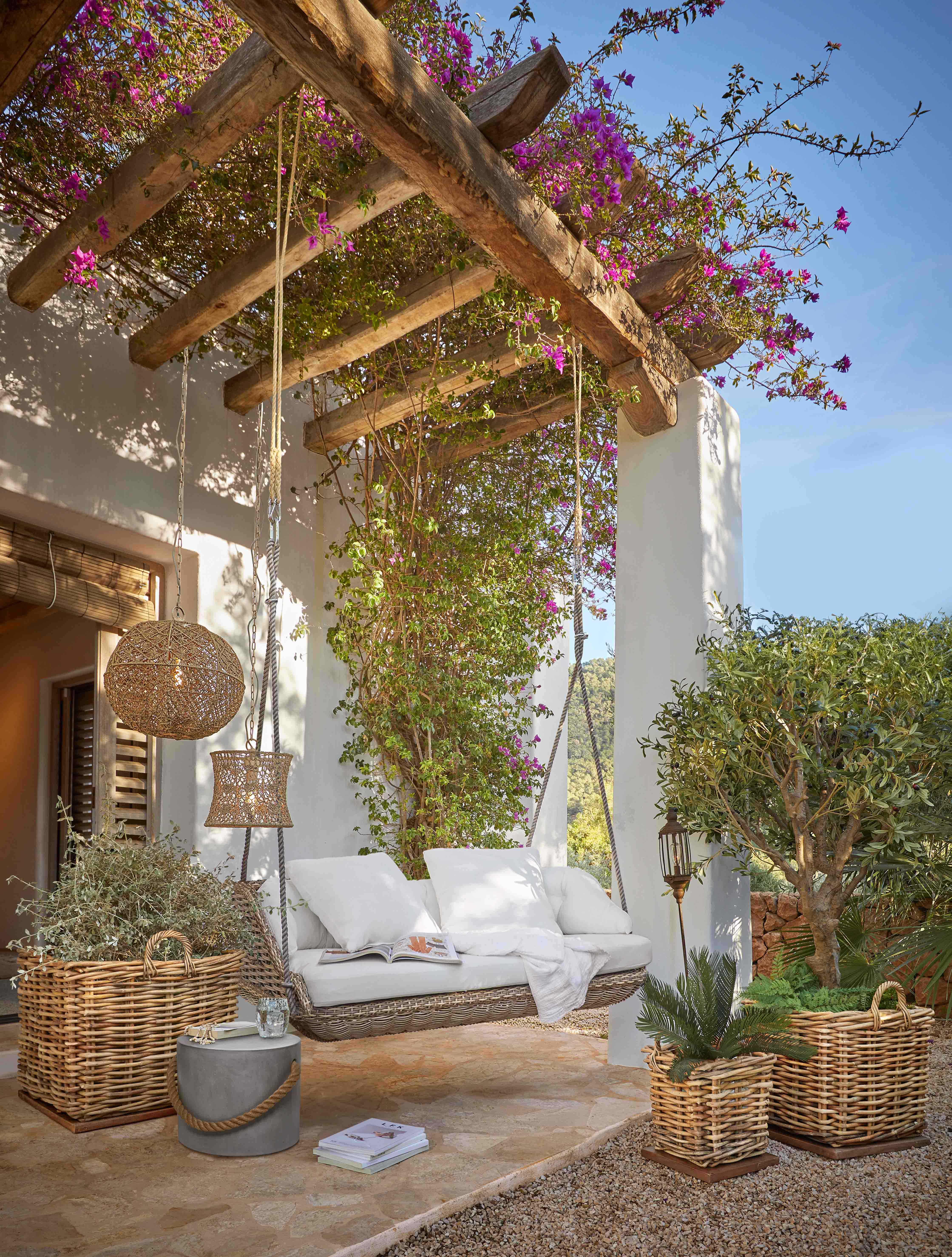 Columpio de mimbre con cojines blancos bajo una pérgola de madera con flores rosas florecientes