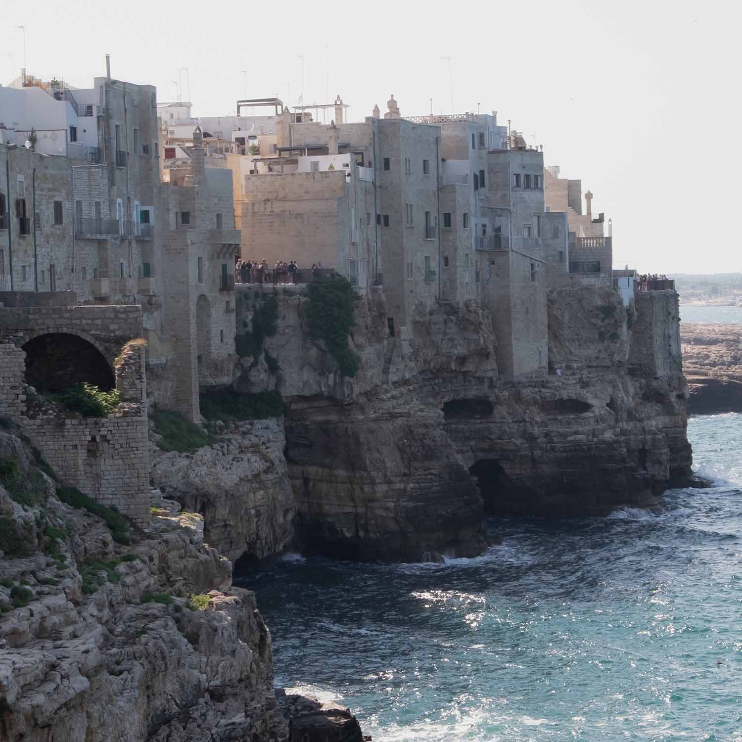 Antiguas casas de piedra en acantilado junto al mar con turistas y olas en la costa