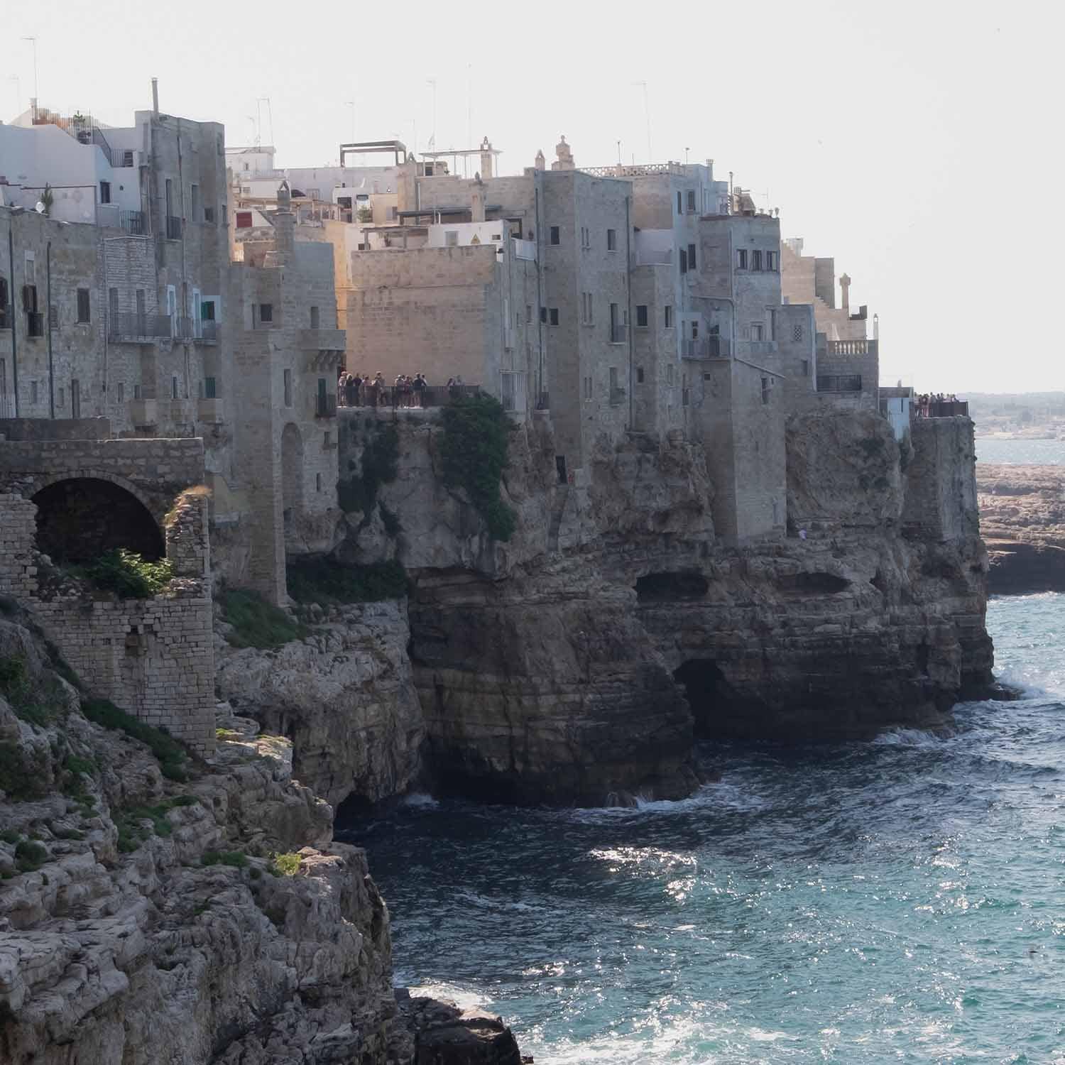 Antiguas casas de piedra en acantilado junto al mar con turistas y olas en la costa