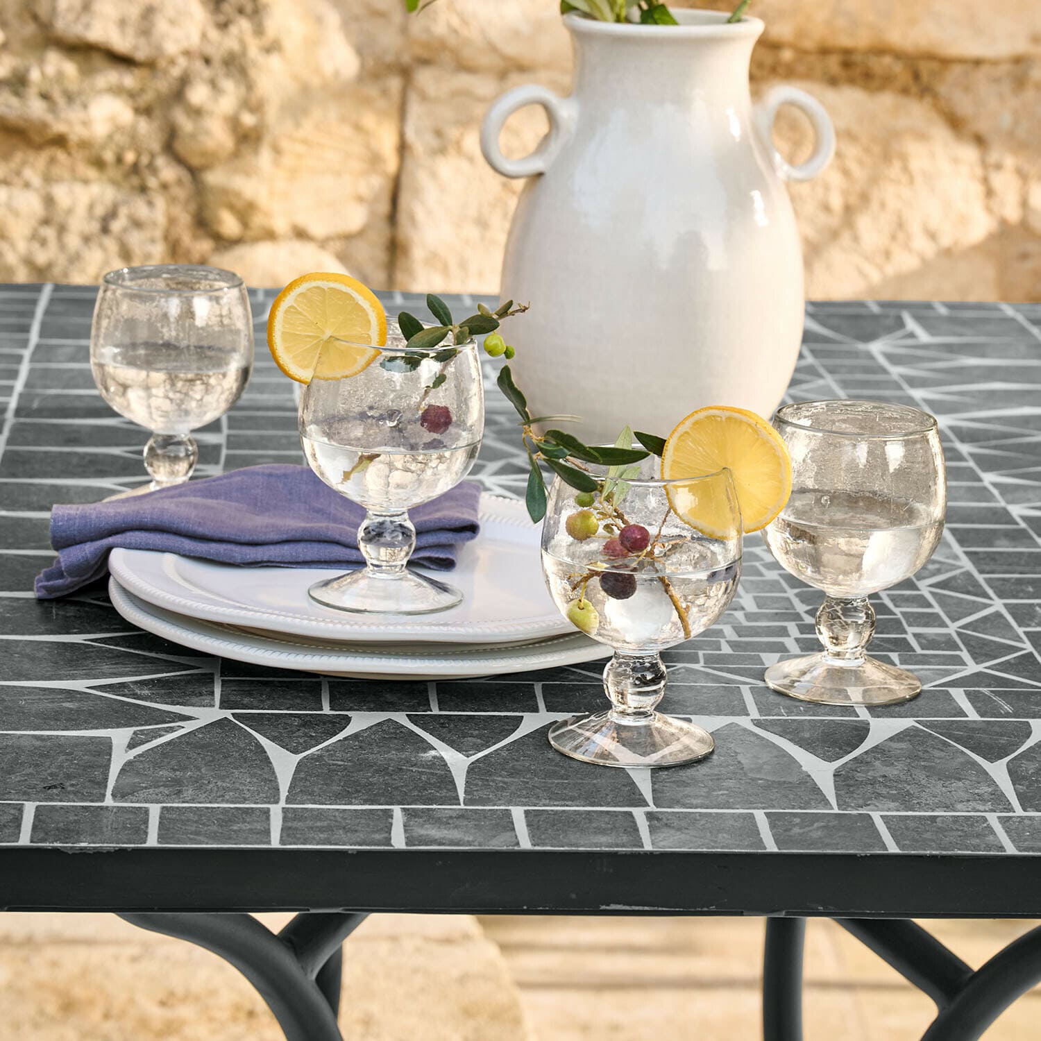 Set de 4 copas Vinoros Copas de cristal con agua y rodajas de limón sobre mesa negra con mosaico, jarrón cerámico blanco al fondo