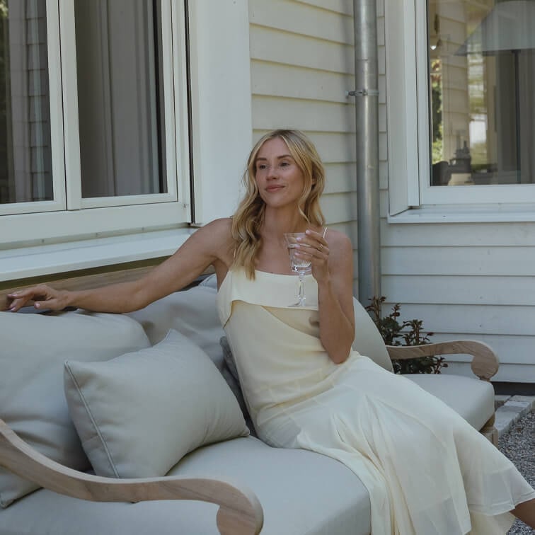 Mujer con vestido crema sentada en sofá gris claro al aire libre, sostiene copa de vino, relajación en terraza
