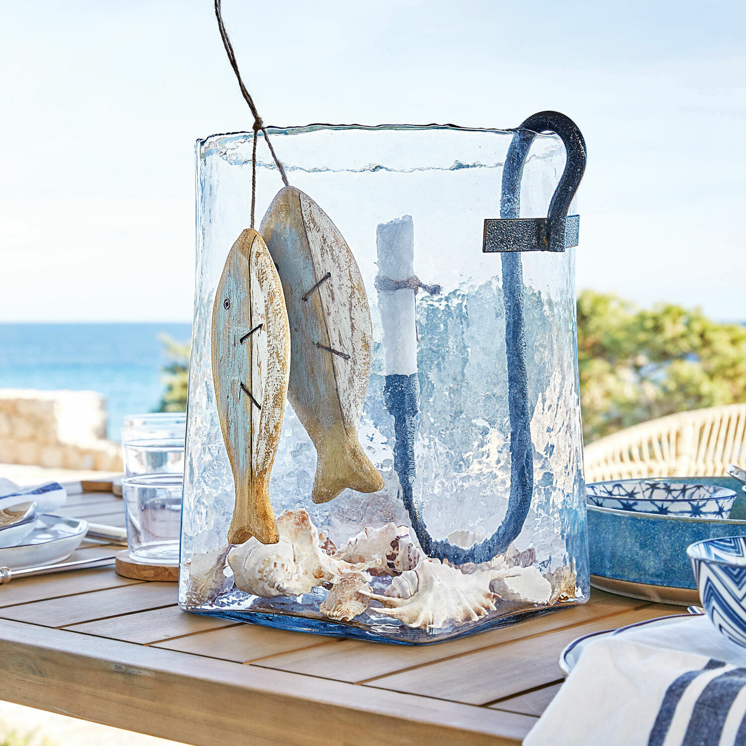 Farolillo Arcene Farol de vidrio con asa de metal, decoración de peces de madera y conchas marinas sobre mesa de madera en terraza