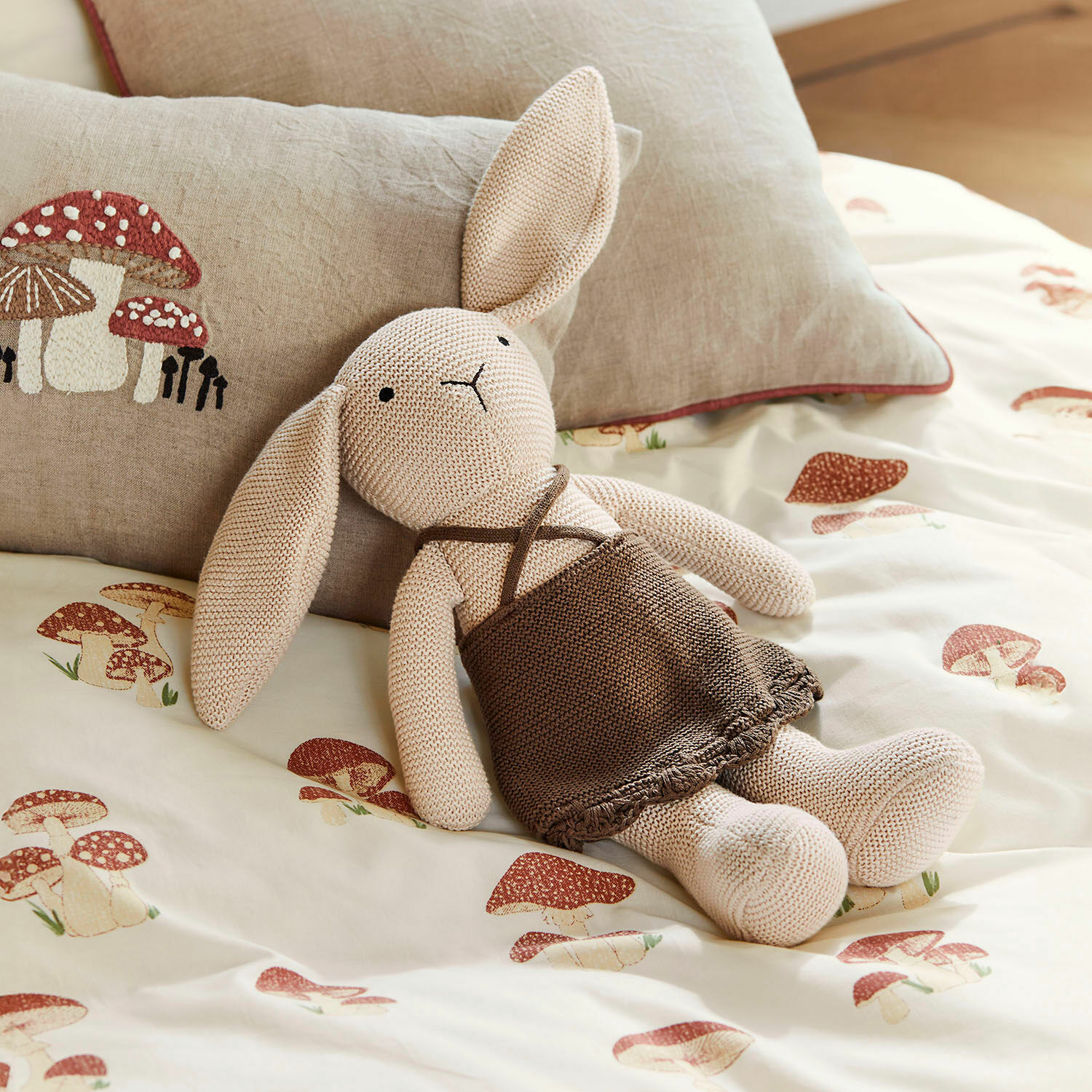 Peluche Flicka Conejo tejido beige con vestido marrón sobre ropa de cama con estampado de setas, cojín decorativo de setas