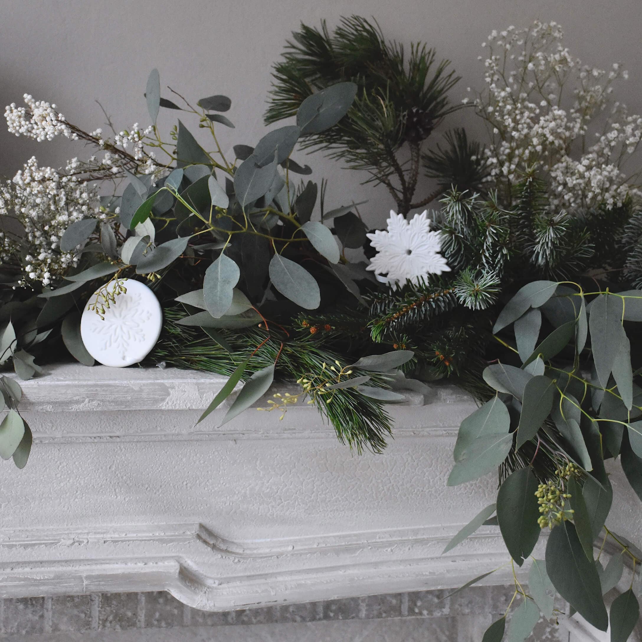 Corona decorativa con ramas verdes y adornos blancos de copos de nieve sobre chimenea blanca de piedra