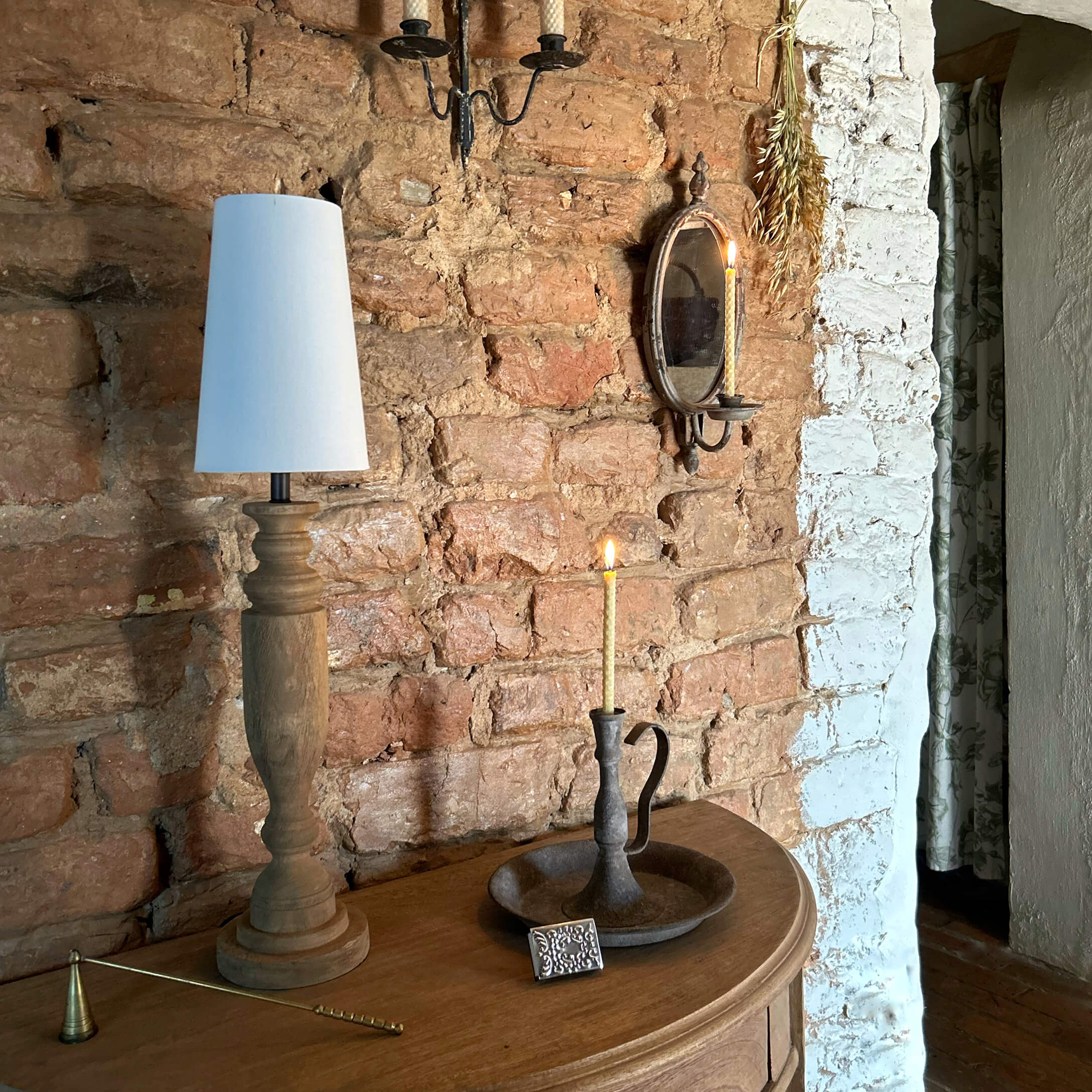 Lámpara de madera con pantalla blanca y candelabro de metal con vela encendida sobre mesa de madera junto a pared de ladrillo