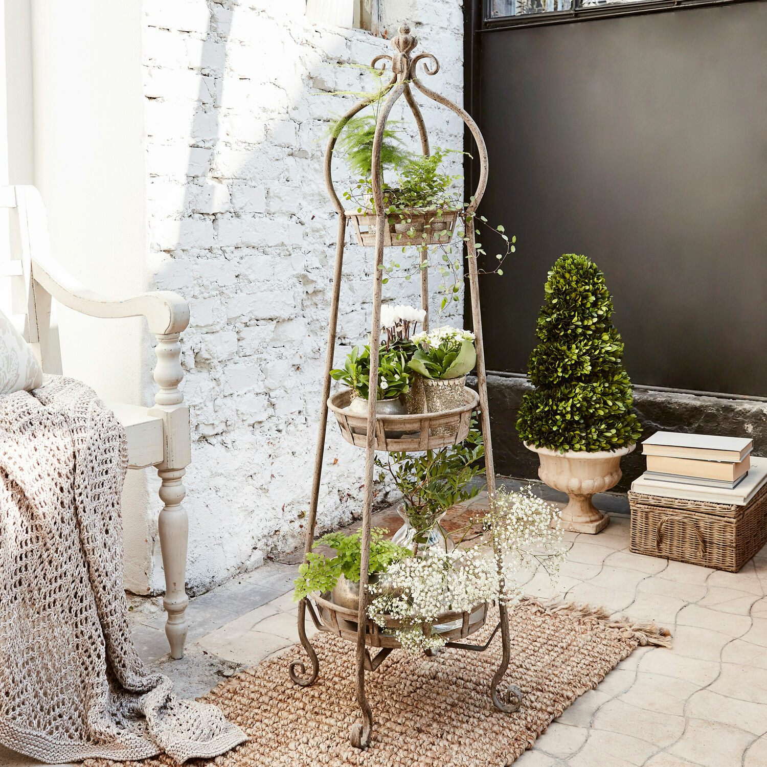 Macetero de metal rústico de pie con tres niveles y plantas, sobre alfombra natural en interior luminoso