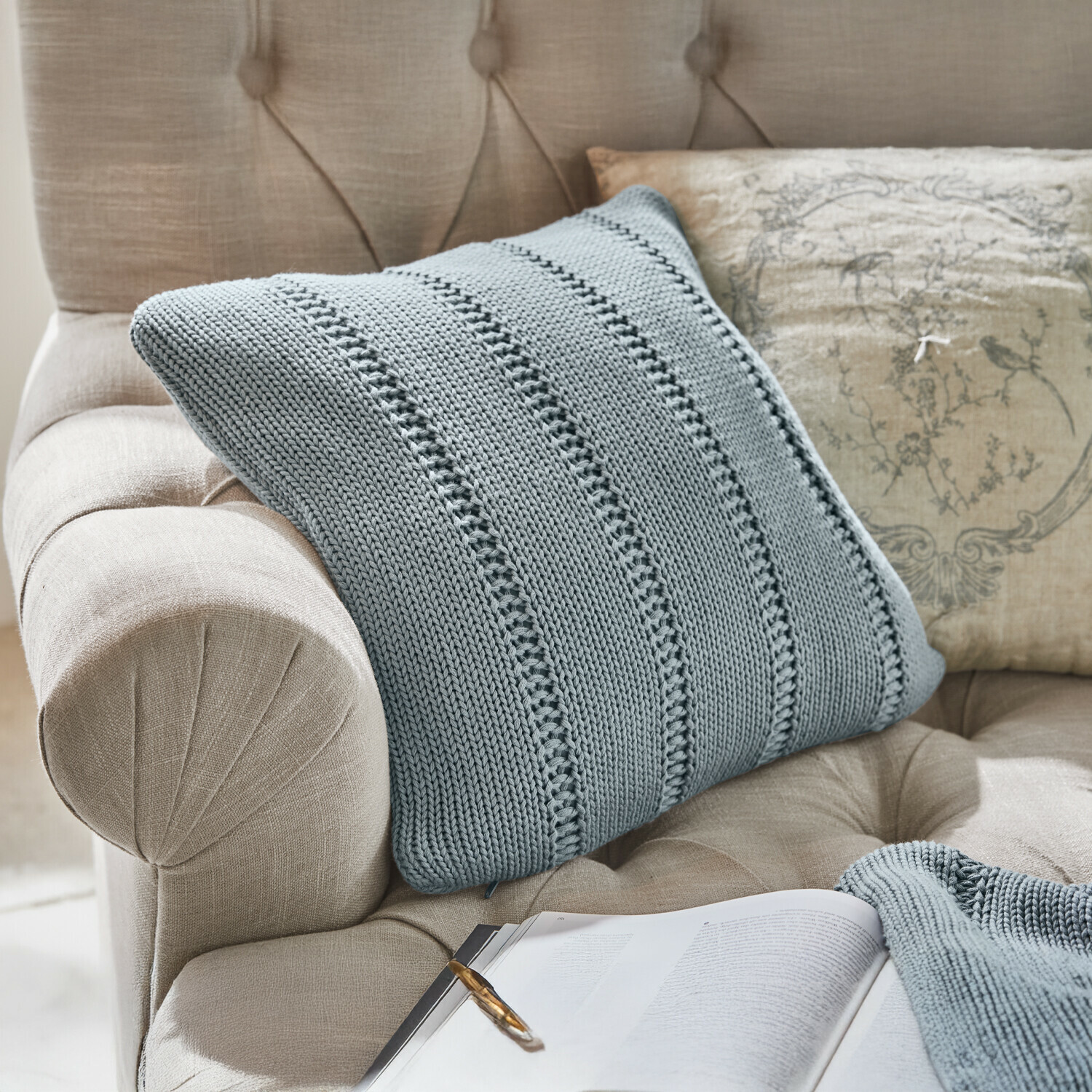 Funda de cojín Lémeré Sillón tapizado beige con respaldo capitoné, manta de punto gris y cojín decorativo
