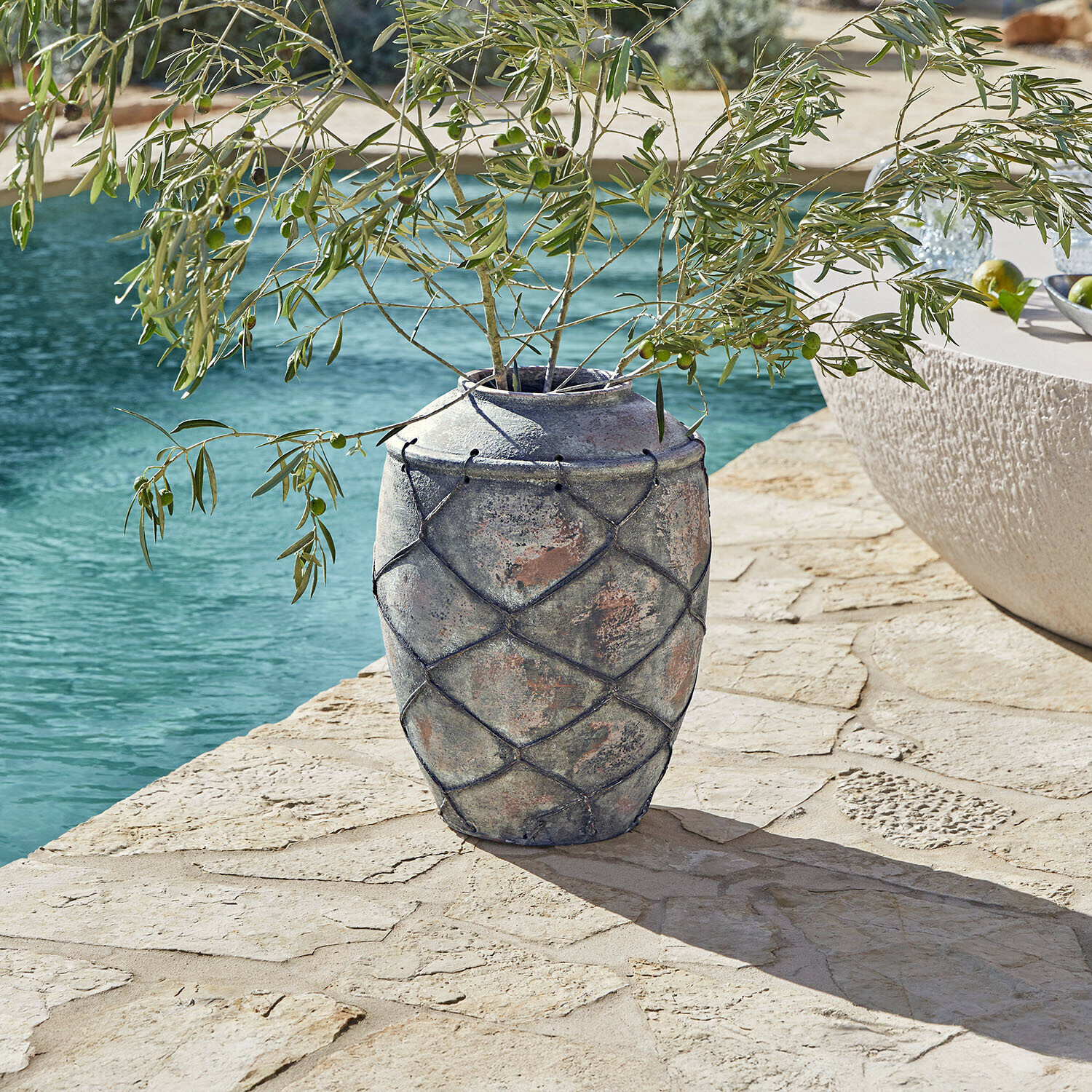 Jarrón Deloriva Jarrón grande de cerámica gris con red decorativa en terraza de piedra junto a piscina con ramas de olivo