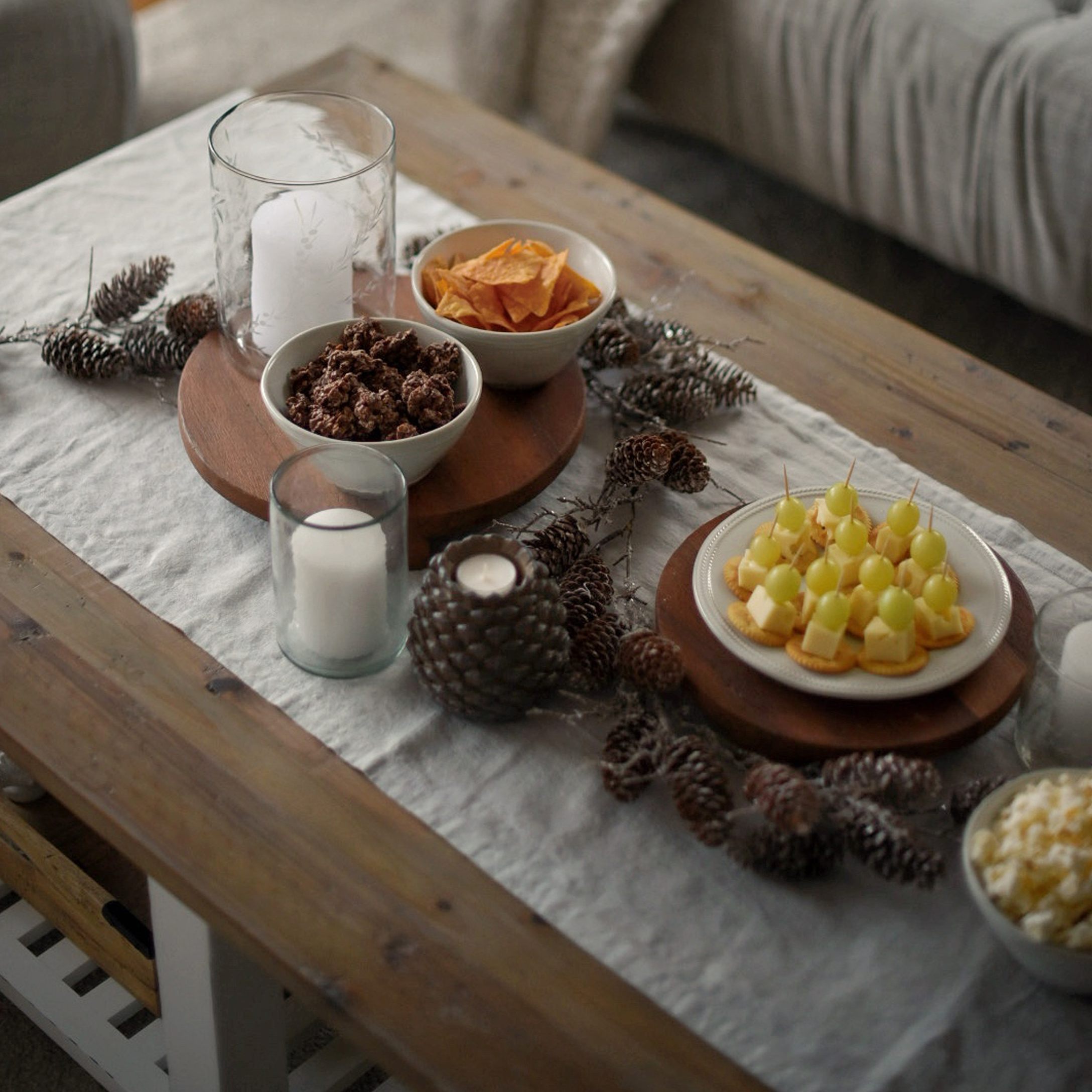 Bandeja de madera con aperitivos y velas sobre mantel de lino con decoración de piñas