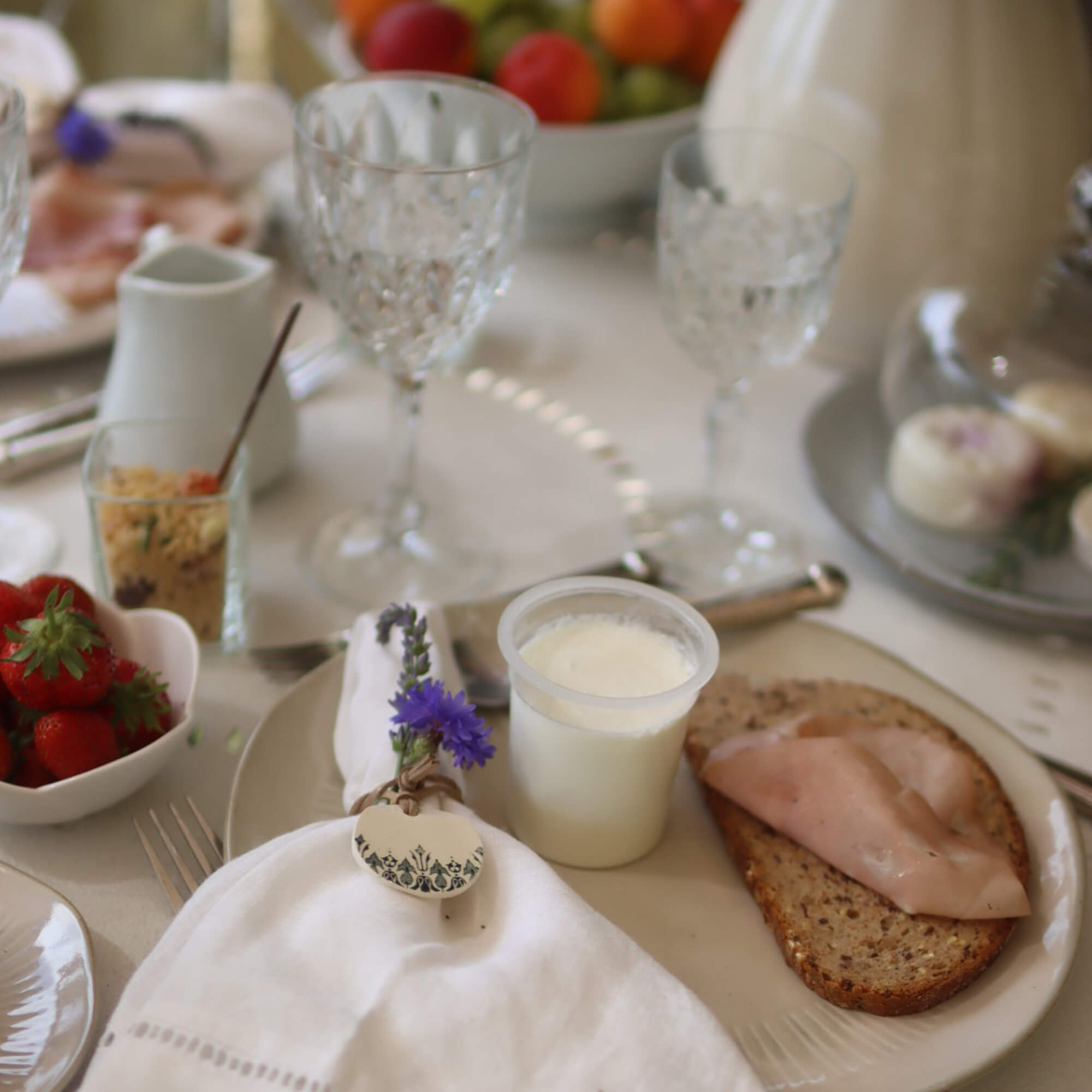 Mesa de desayuno con pan integral, jamón y yogur en plato, servilleta decorada con flores
