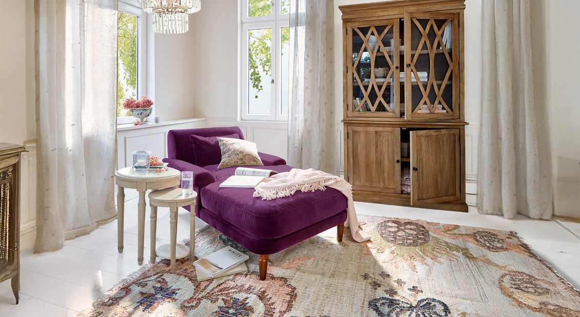 Chaise longue de terciopelo morado con cojines y manta en salón luminoso con armario de madera y alfombra