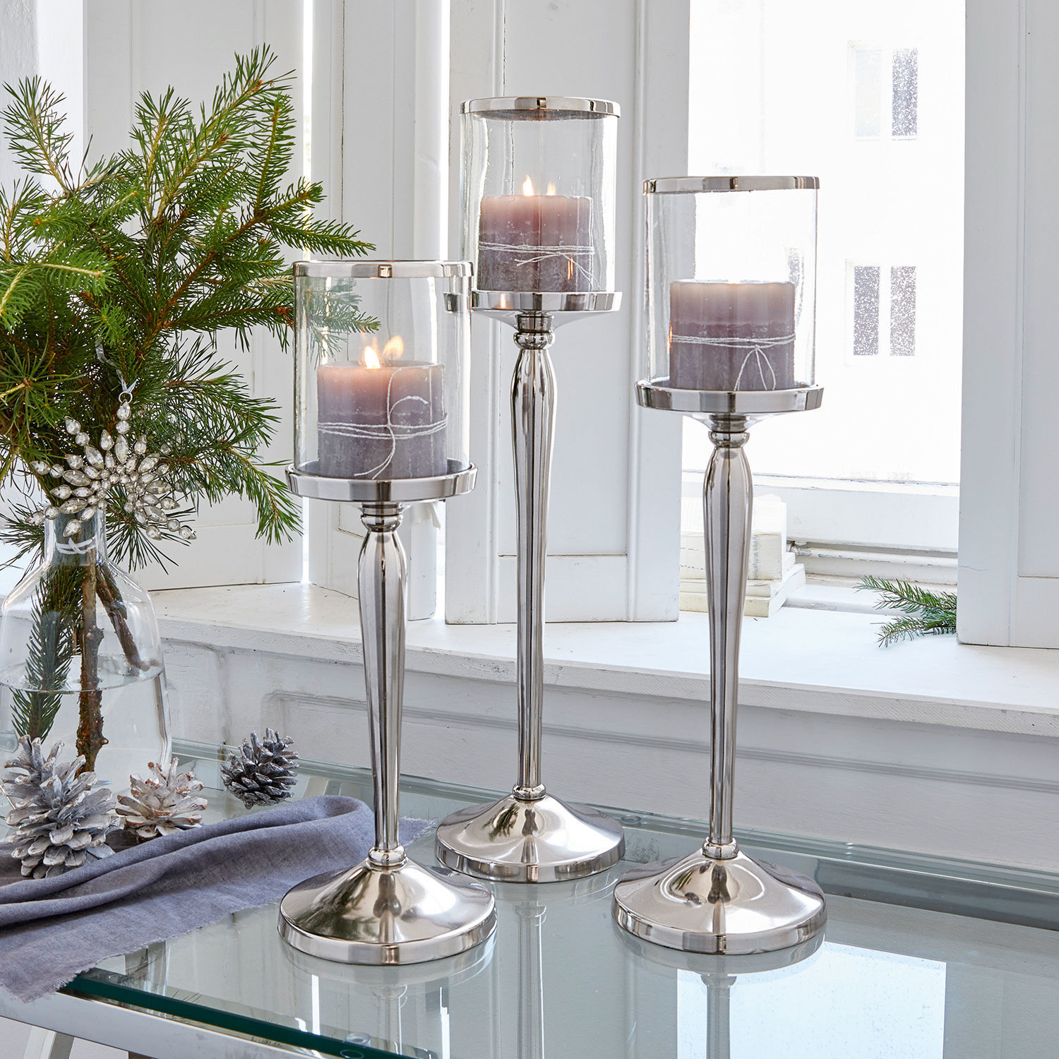 Juego de 2 candelabros Serres Conjunto de tres candelabros plateados de vidrio y metal con velas moradas sobre mesa de cristal