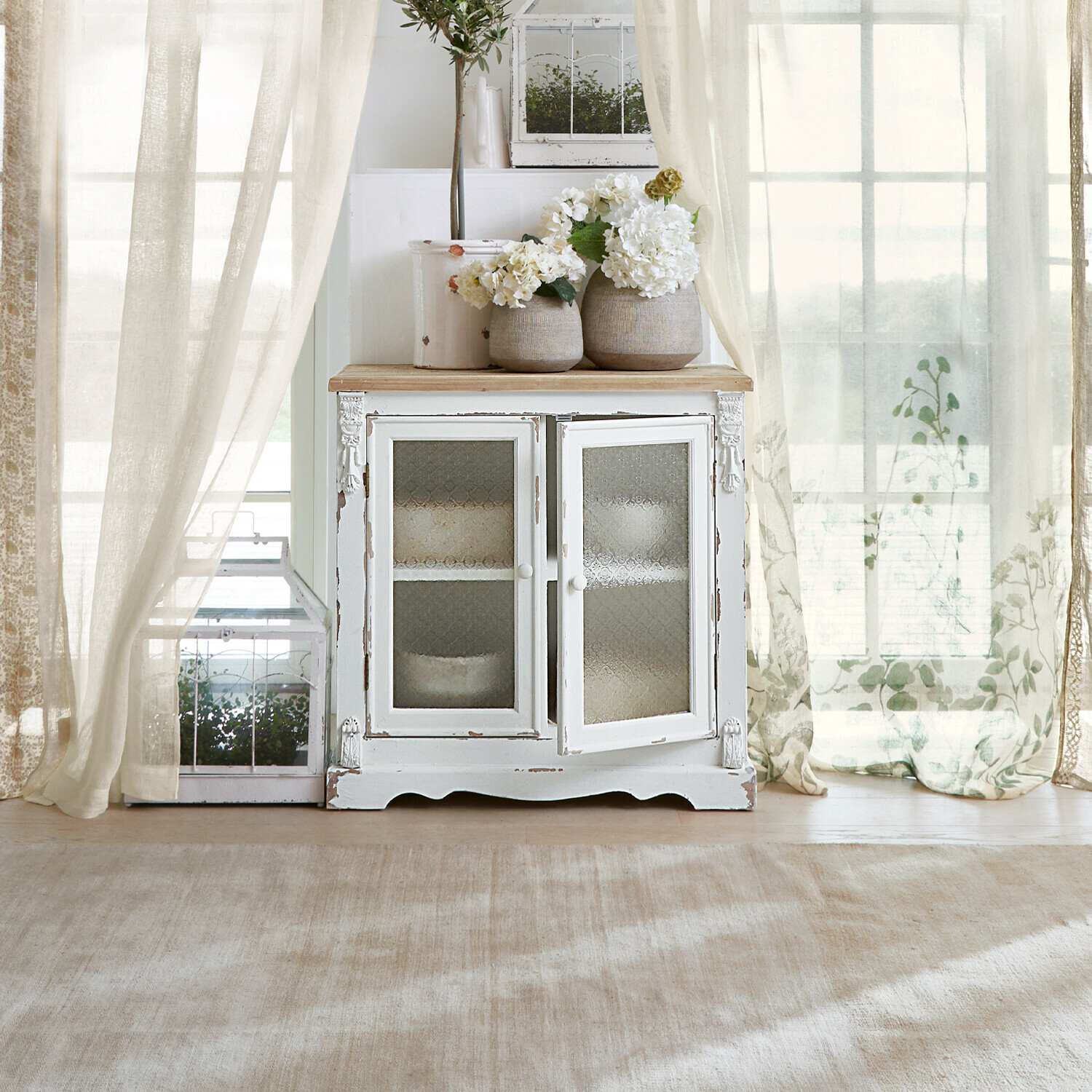 Cómoda South Yarmouth Aparador de madera blanca con puertas de cristal y jarrones decorativos con flores en interior luminoso
