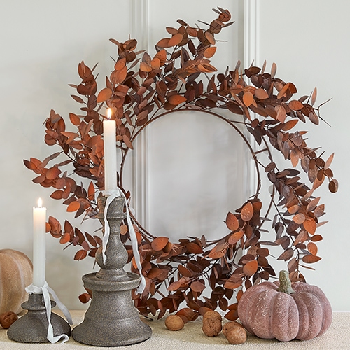 Corona decorativa de hojas metálicas oxidadas con dos velas y calabaza decorativa sobre mesa