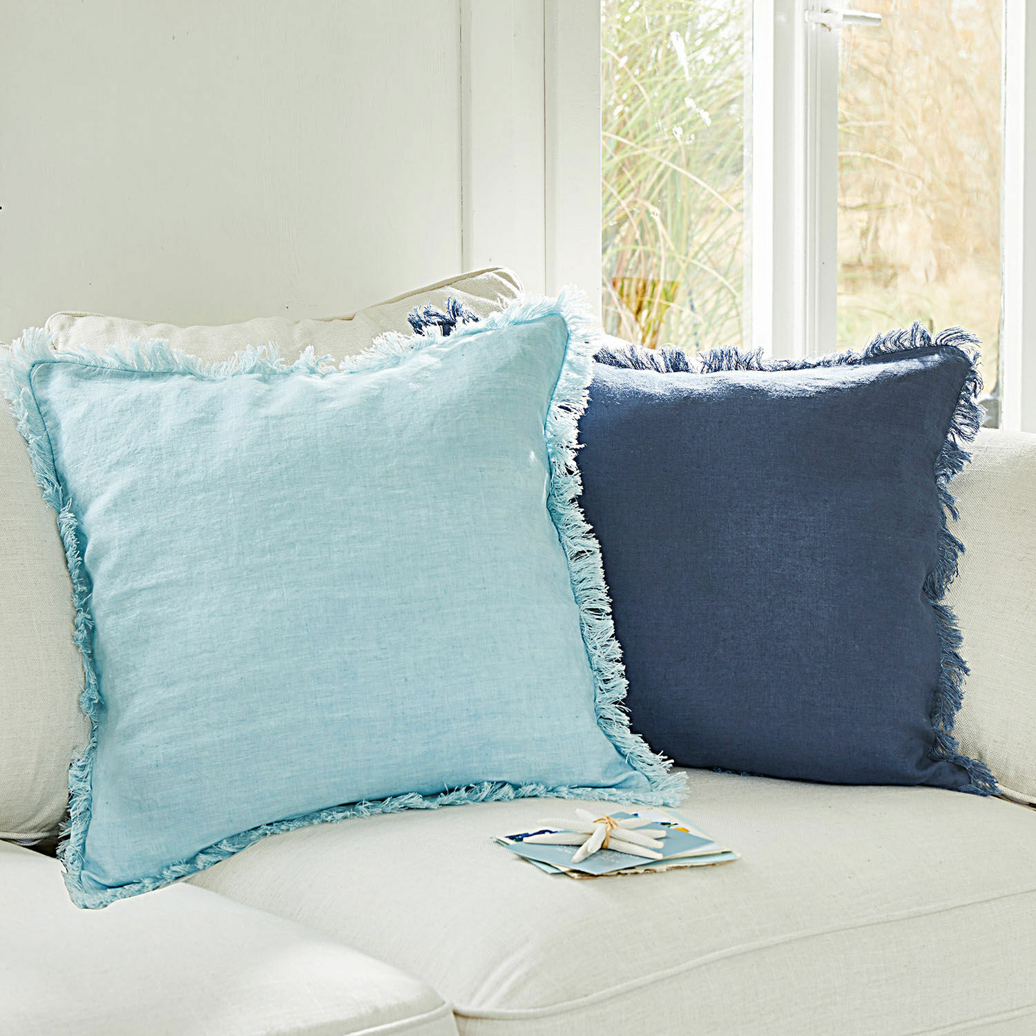 Set de 2 fundas de cojín Quebo Conjunto de dos cojines decorativos de lino azul claro y azul marino con flecos sobre un sofá beige