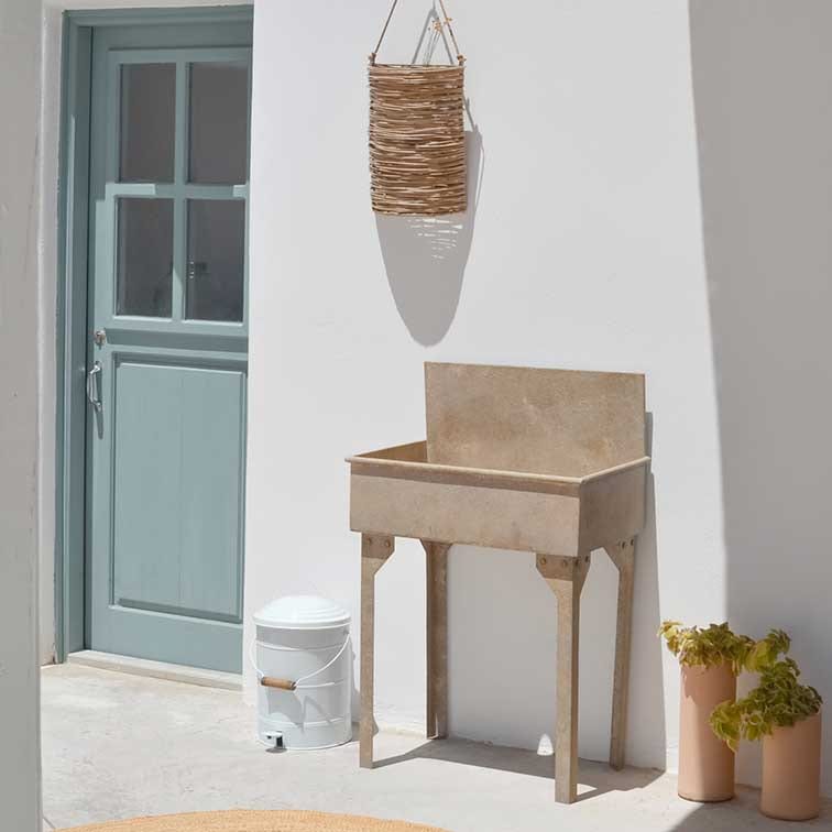 Maceta beige de piedra con patas junto a pared blanca, puerta azul y cesta de mimbre colgada