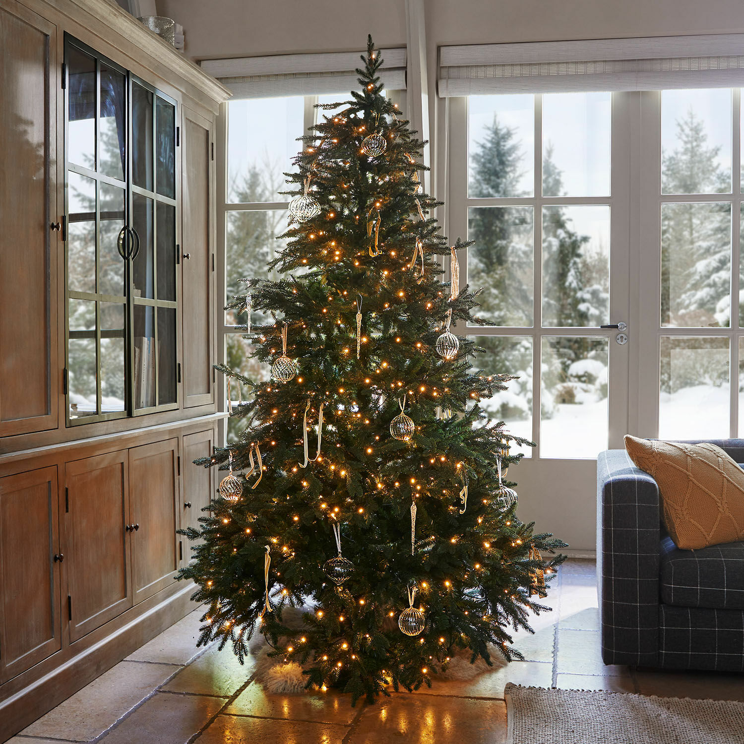 Árbol de Navidad Celindor Árbol de Navidad iluminado con ramas verdes y adornos de vidrio en sala acogedora con vista a la nieve