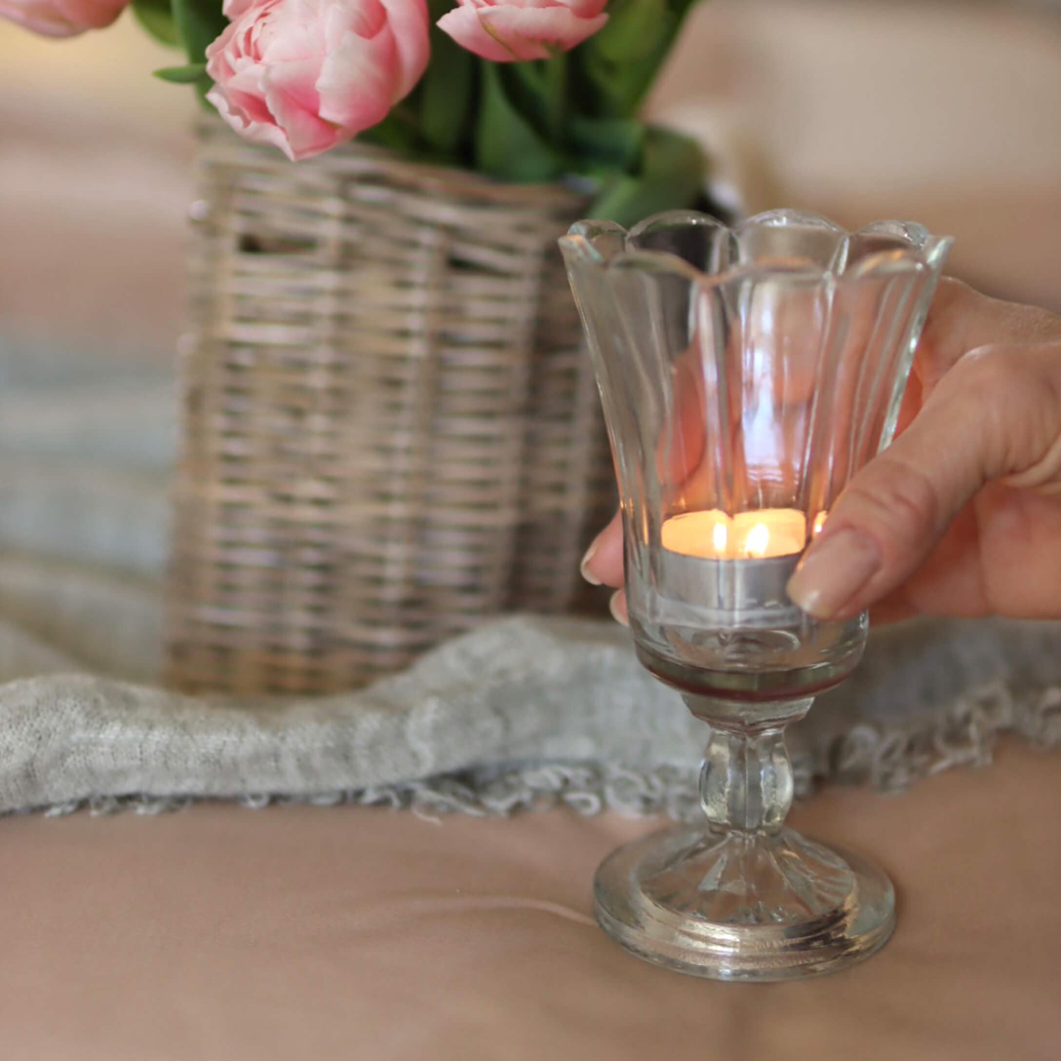Portavelas de vidrio con vela encendida, jarrón con flores rosas y cesta de mimbre al fondo