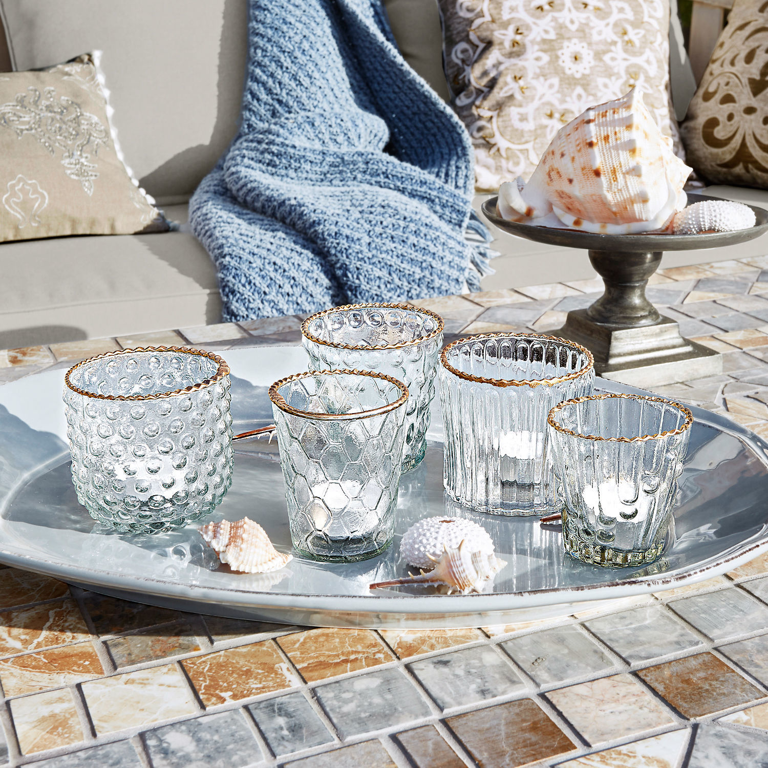 Set de 3 farolillos Jarnac Conjunto de cinco portavelas de vidrio con borde dorado sobre bandeja plateada en mesa de mosaico