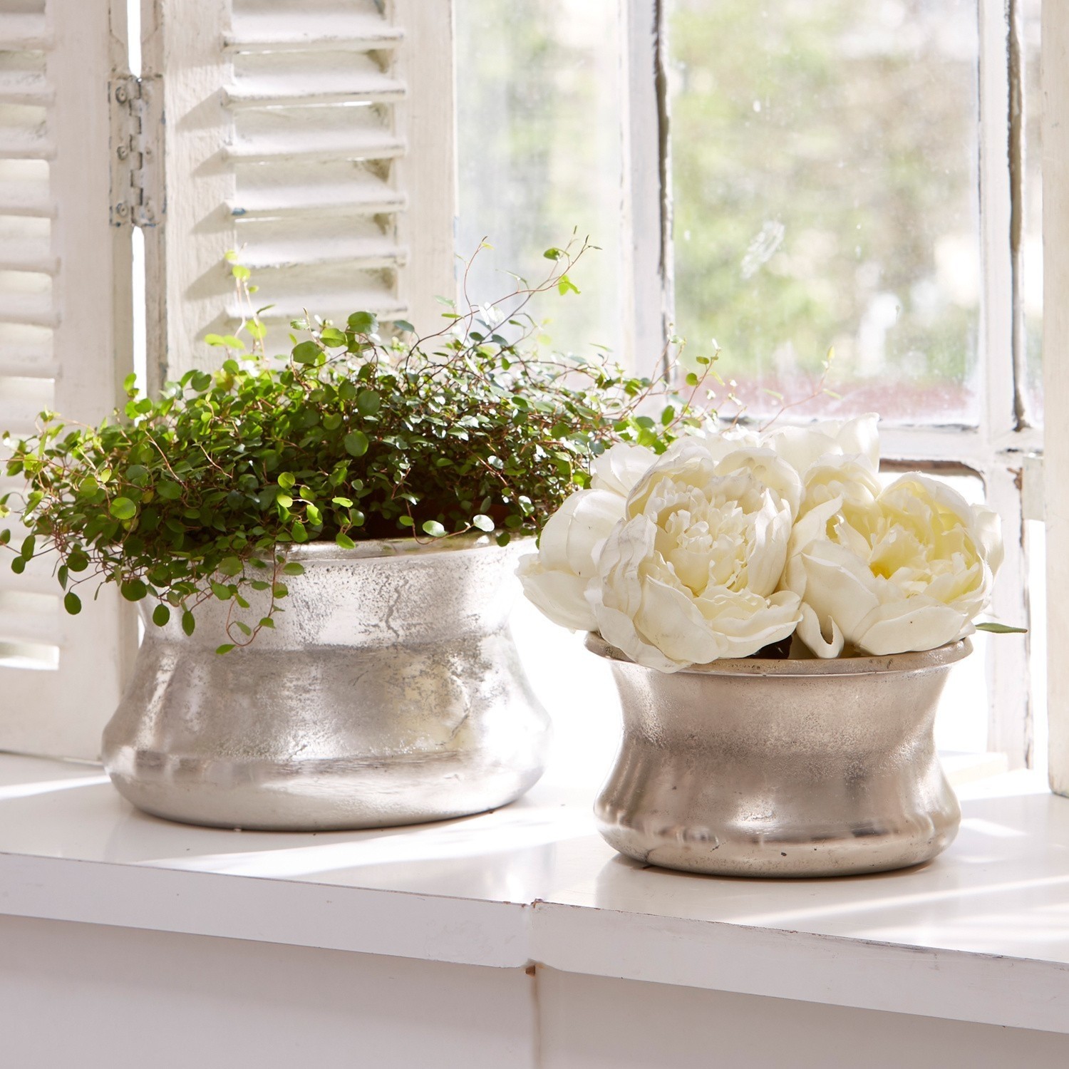 Set de 2 jarrones Riona Dos macetas metálicas plateadas en el alféizar con planta verde y flores blancas decorativas