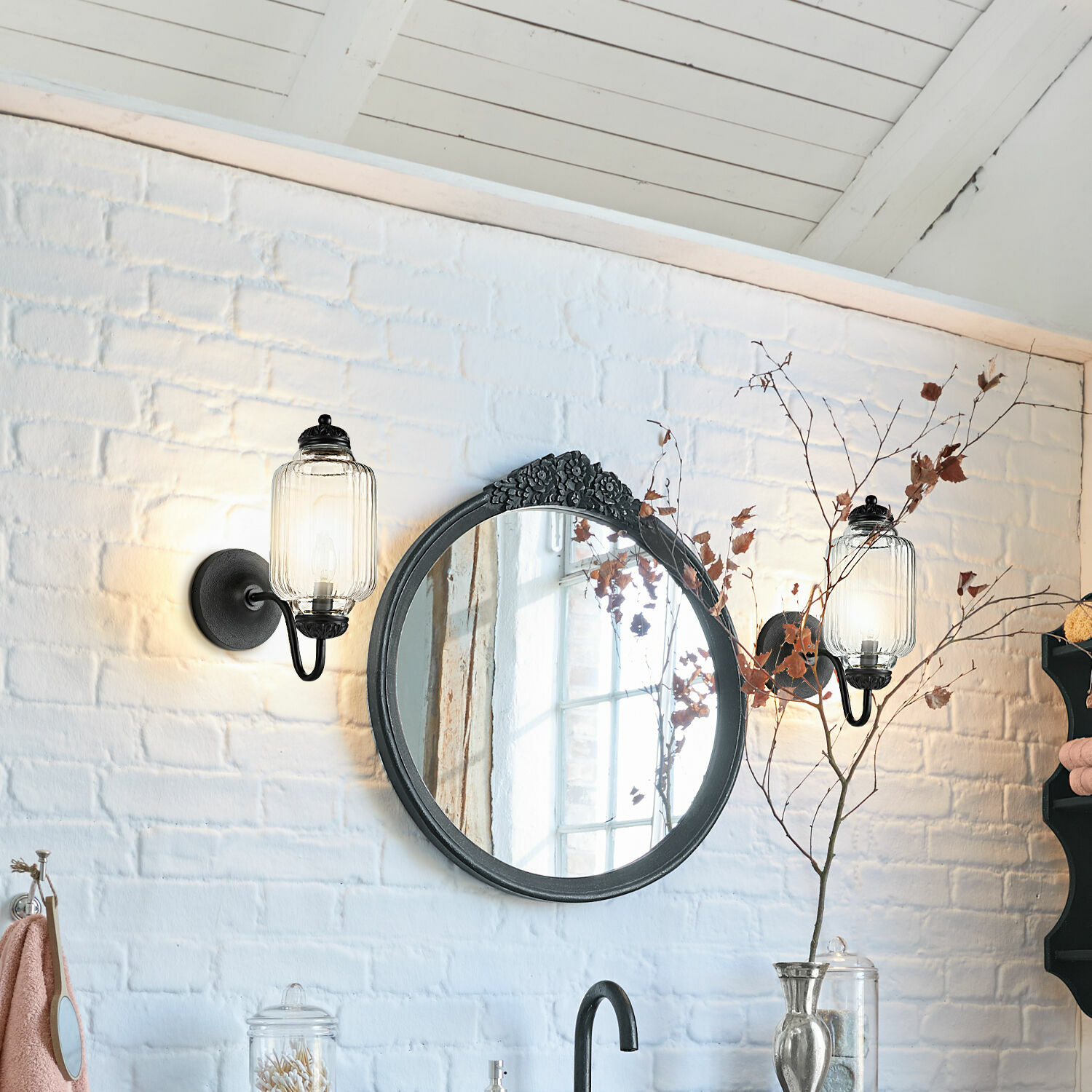Espejo redondo con marco negro y decoración floral en pared de ladrillo blanco con apliques de pared