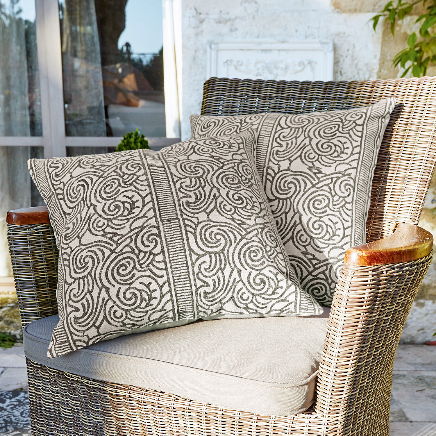 Set de 2 fundas de cojín Ensis Dos cojines decorativos de algodón beige con patrón marrón en sillón de mimbre en terraza