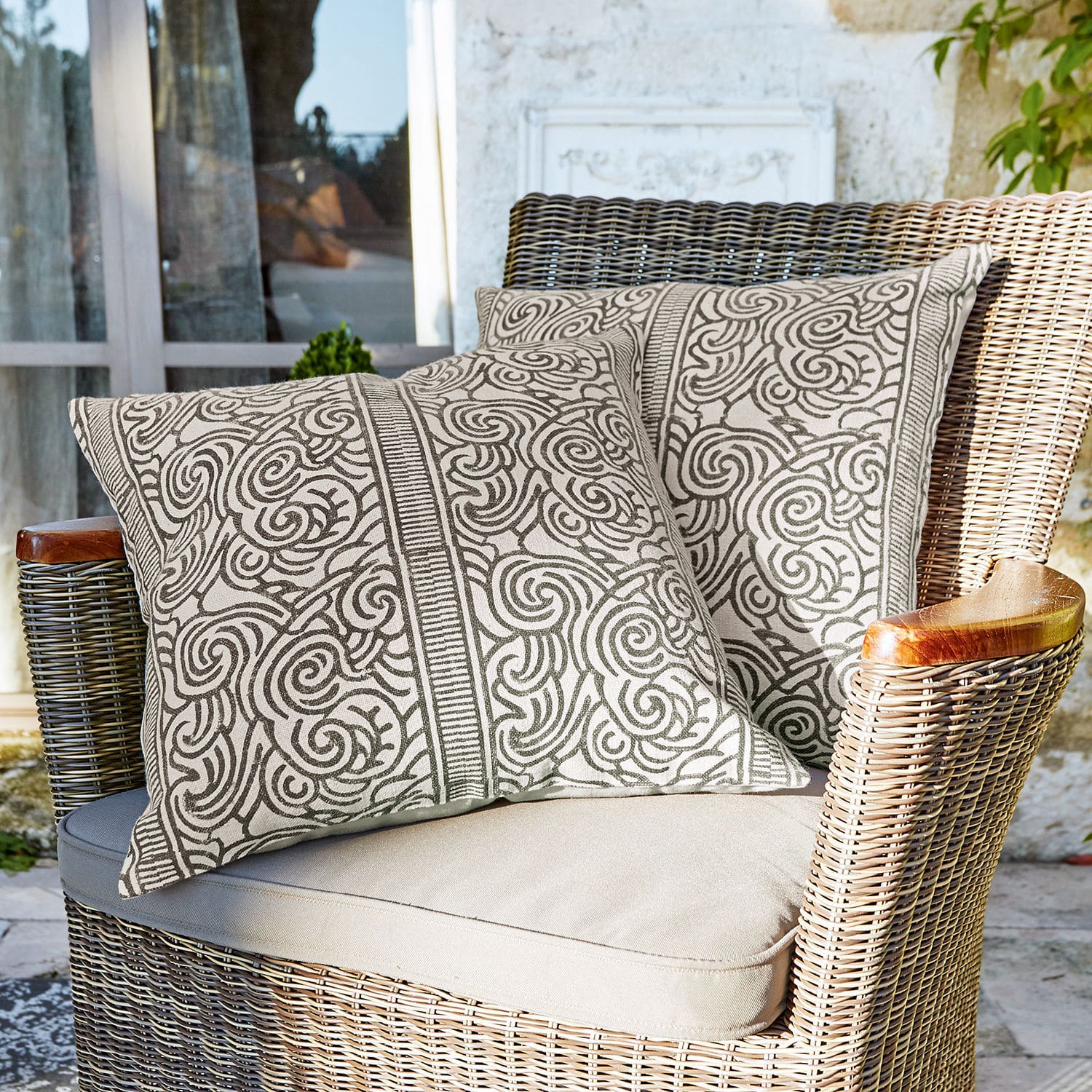 Set de 2 fundas de cojín Ensis Dos cojines decorativos de algodón beige con patrón marrón en sillón de mimbre en terraza