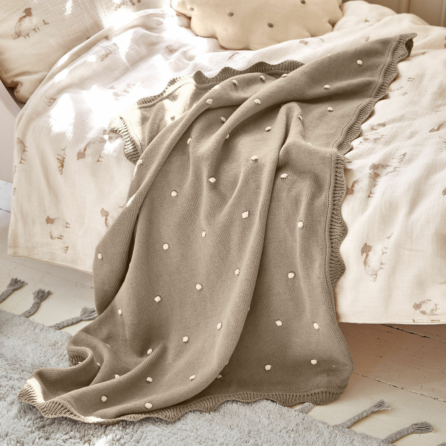 Plaid Saphie Manta beige de punto con lunares blancos sobre cama con ropa clara, suave y decorativa