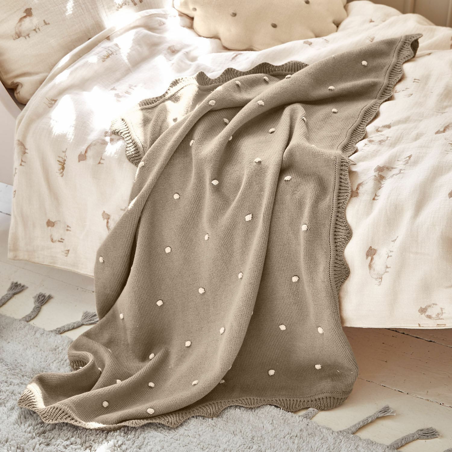 Plaid Saphie Manta beige de punto con lunares blancos sobre cama con ropa clara, suave y decorativa
