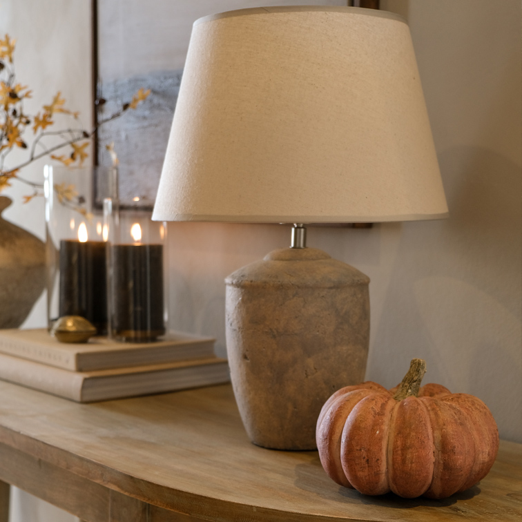 Lámpara de mesa con pantalla beige y base de piedra sobre mesa de madera con decoración de calabaza