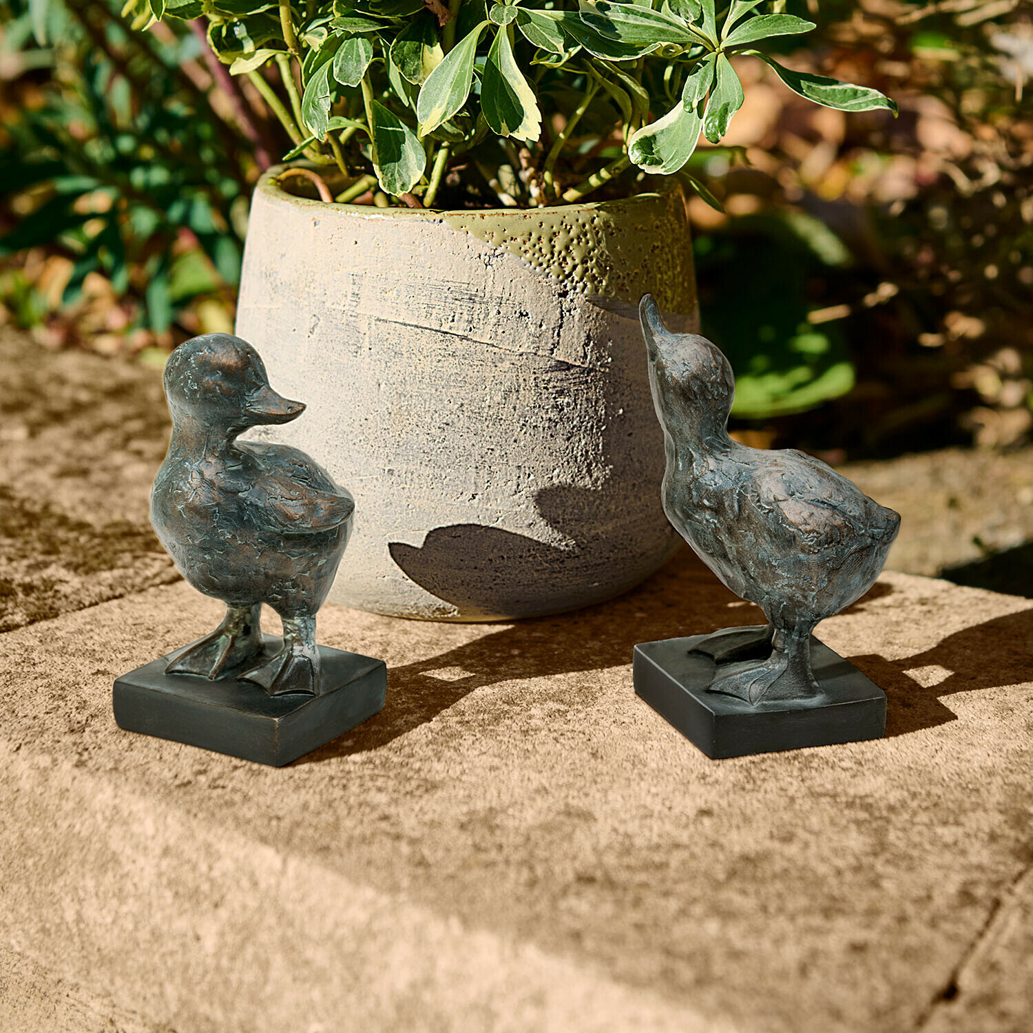 Dos figuras decorativas de patos de metal oscuro sobre superficie de piedra junto a una maceta